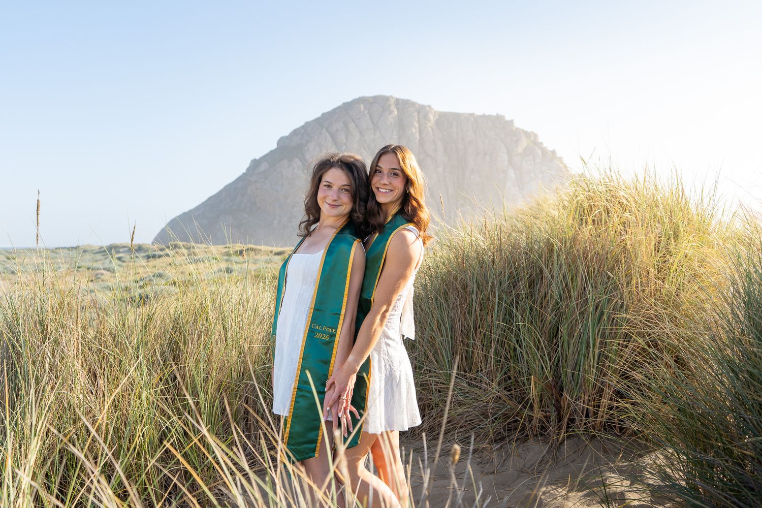Two women pose in sand dunes near Morro Rock, one wearing a green graduation stole, on a sunny day.
