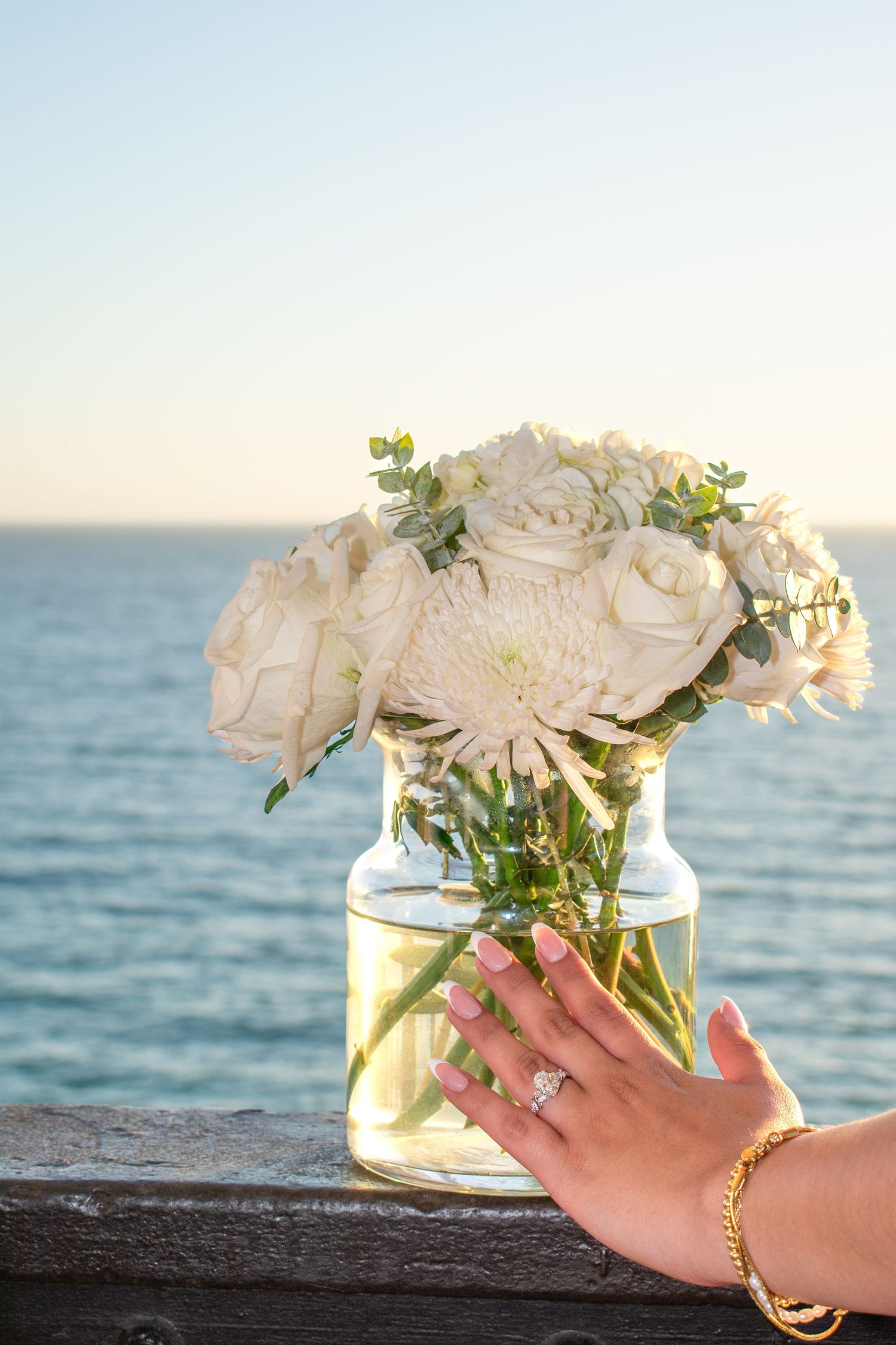 White roses in a glass vase displayed on wooden railing with ocean backdrop.