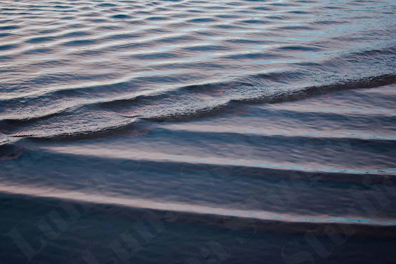 Gentle rippling waves on calm water surface at dusk, with soft blue and pink hues reflecting serene coastal scenery.