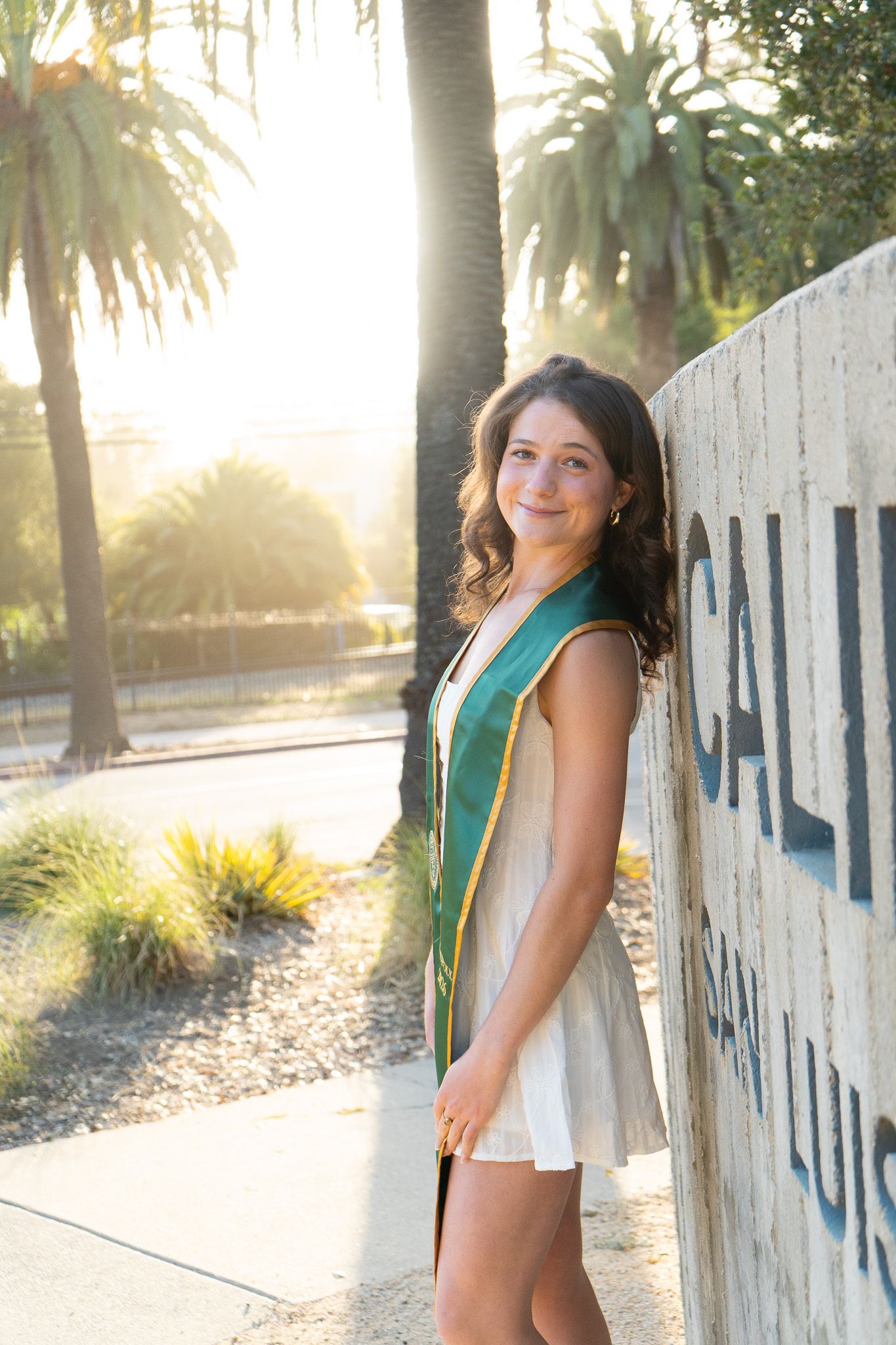 A smiling graduate poses in a white dress and green sash next to a Cal Poly San Luis Obispo sign surrounded by palm trees.