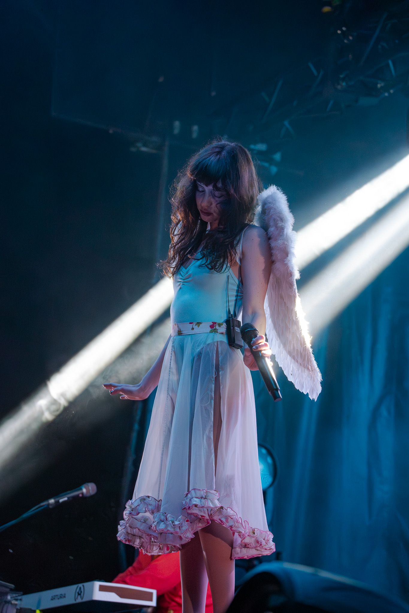 Woman in angel costume with white wings and sheer dress performing on stage under dramatic spotlights.