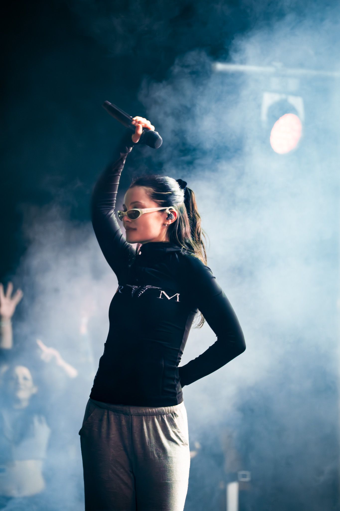 Female performer holds microphone above head on smoky stage, wearing black top and sunglasses under dramatic concert lighting.