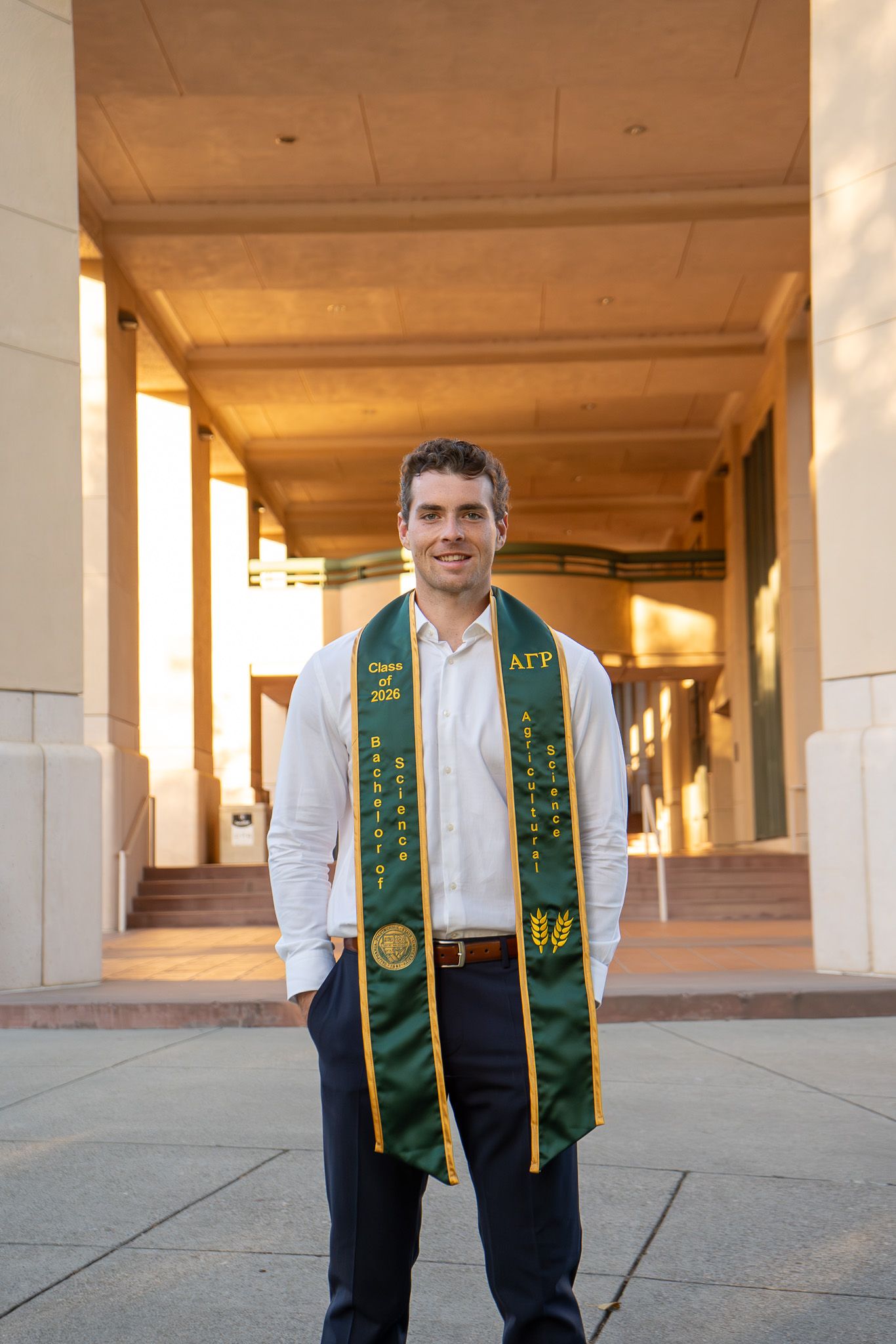 Young man in white shirt wearing green AGP graduation stole smiles outdoors at university building.