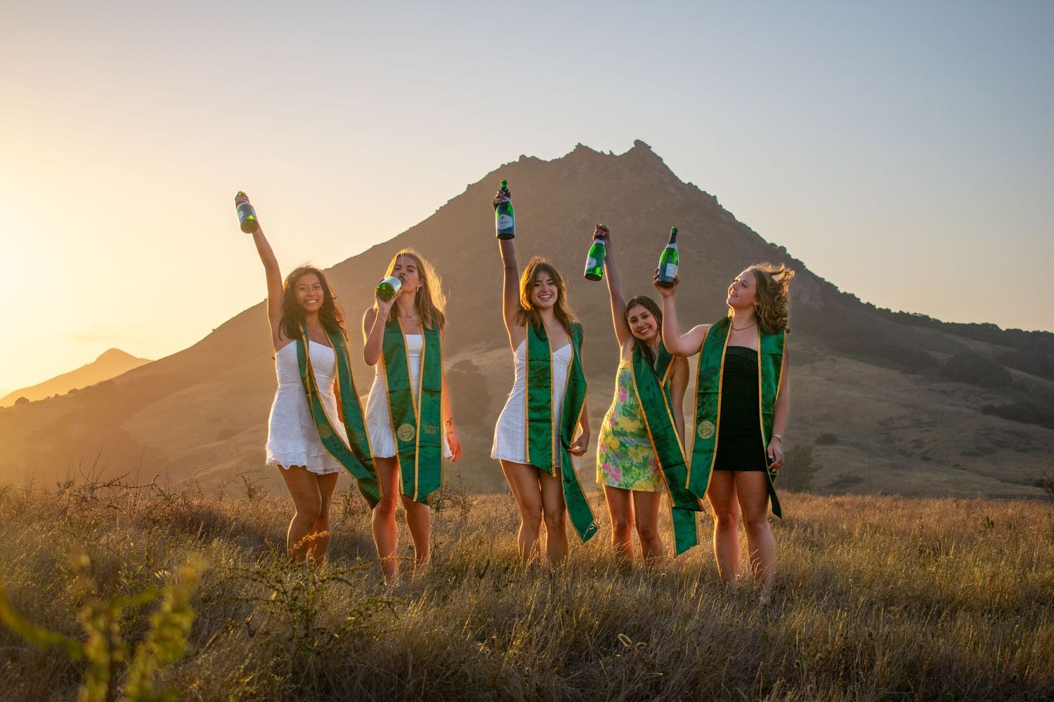 Group of graduates pose with raised arms against mountain backdrop at sunset.