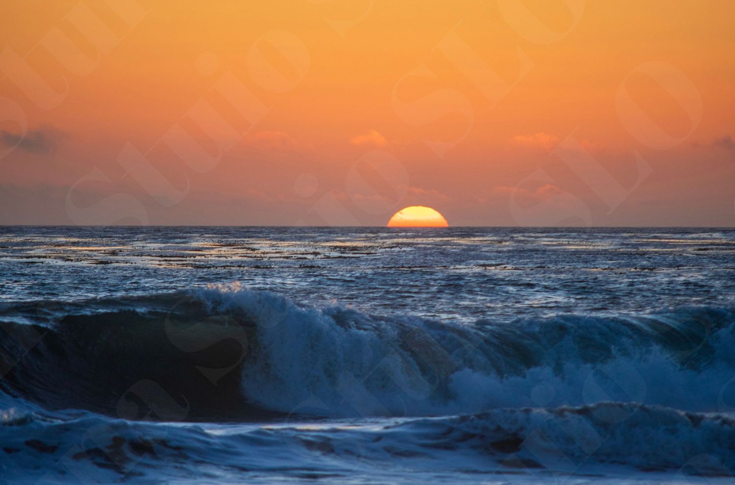 Ocean waves roll in during a dramatic orange sunset with the sun dipping below the horizon.