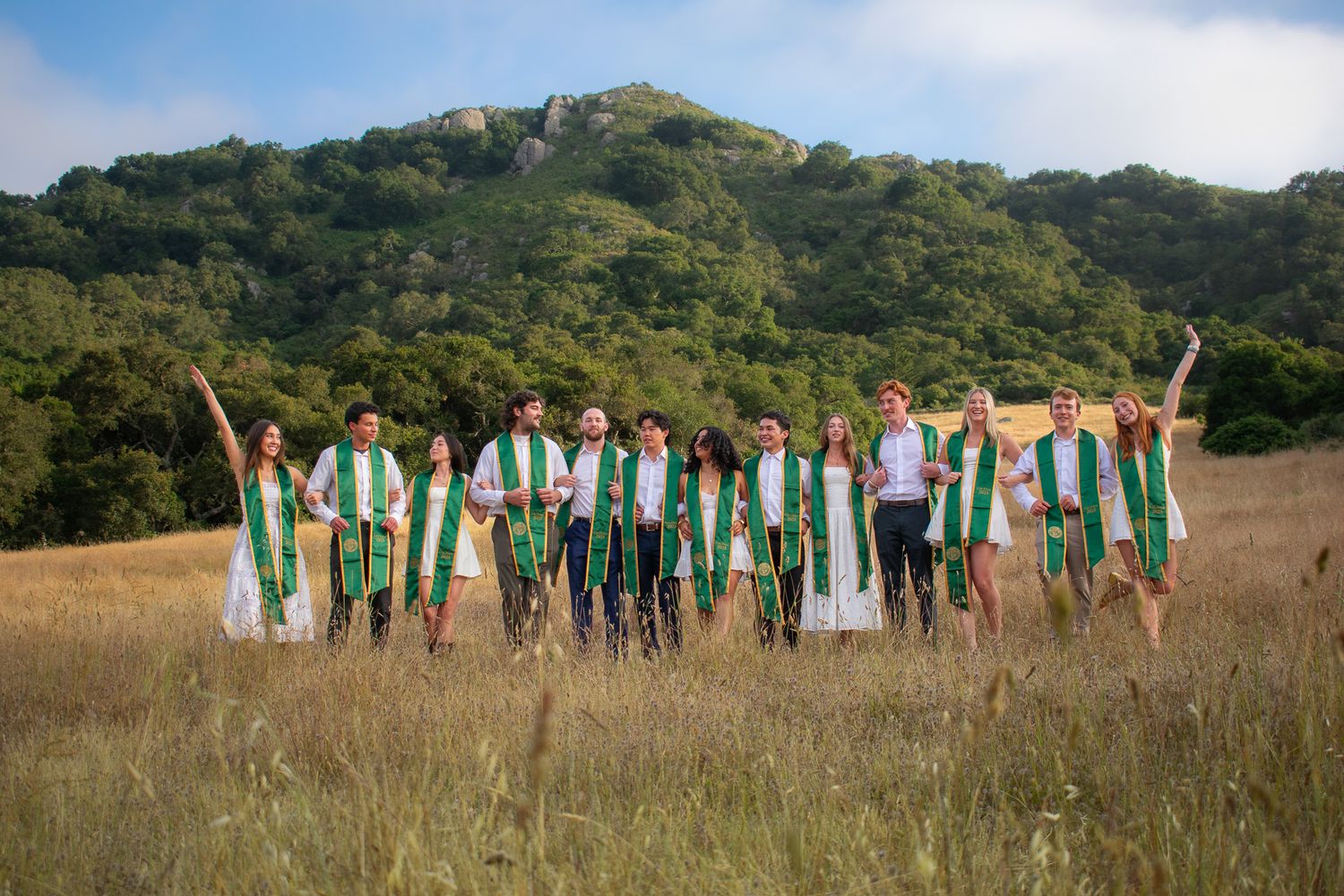 Large group of graduates in green and white pose energetically across grassy hillside.