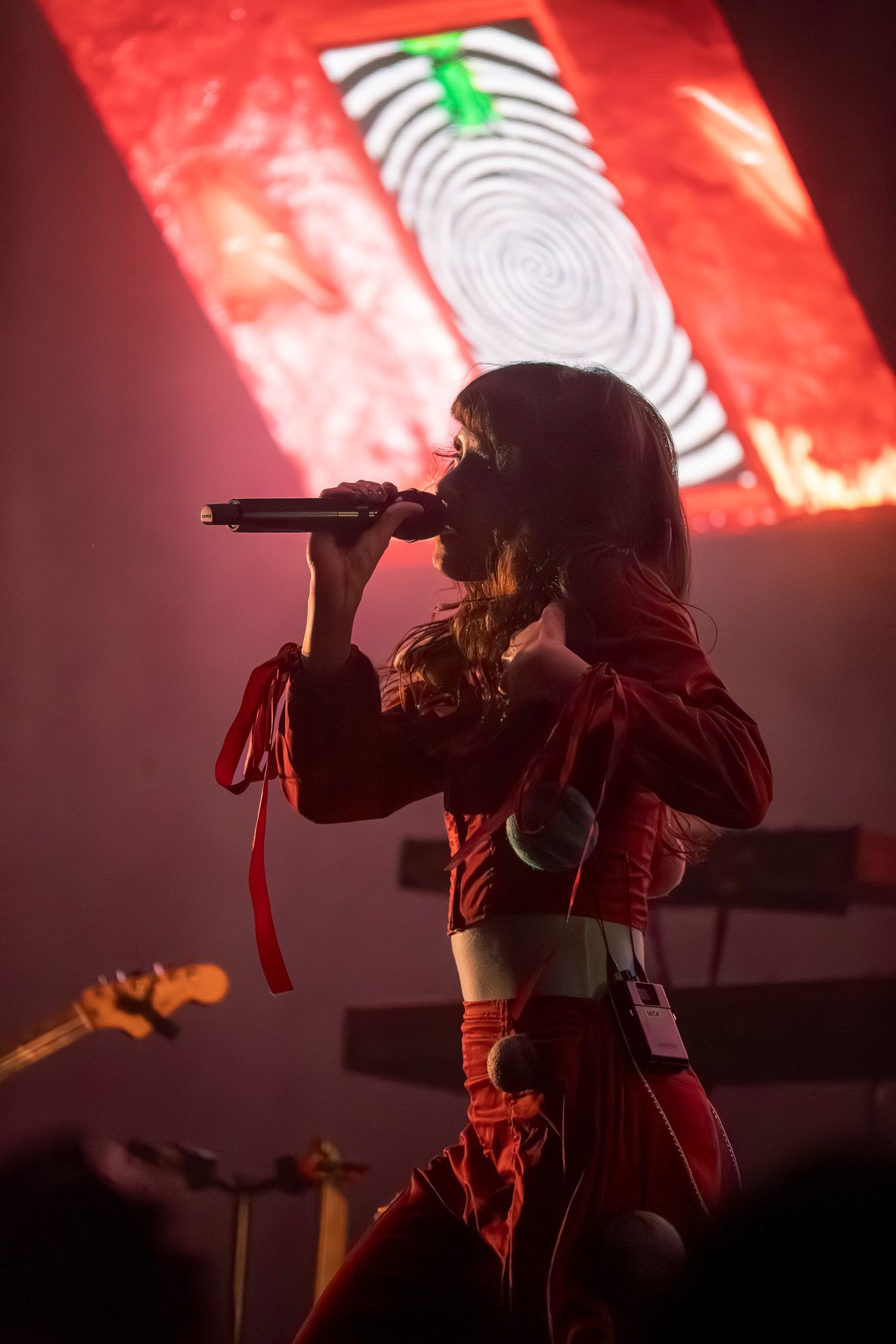 Female singer performing on stage in red outfit, holding microphone with red light backdrop at live concert.