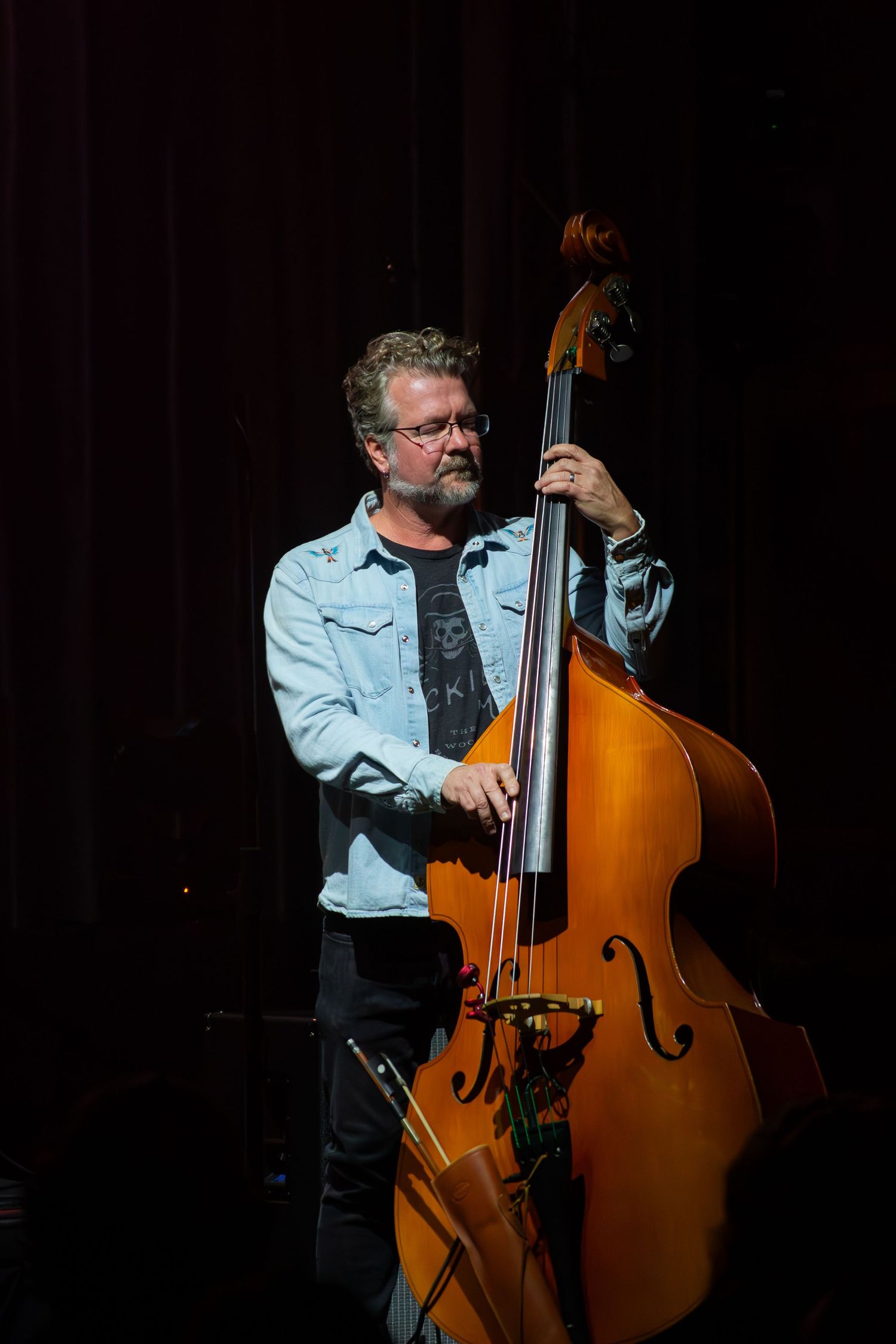 Double bass player performs on dark stage in denim jacket.