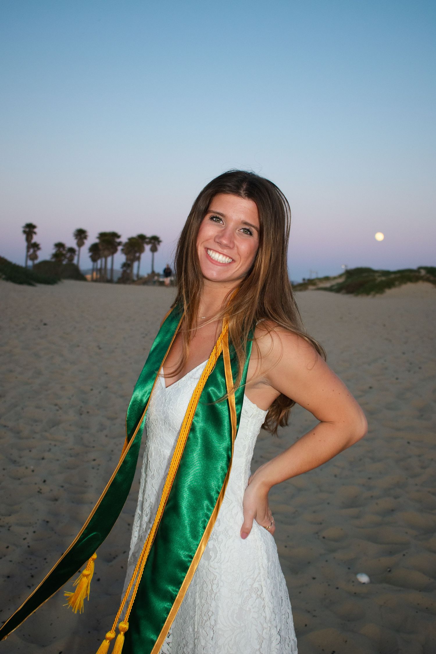 Graduate in white dress and green sash poses for sunset photos on a beach with palm trees.