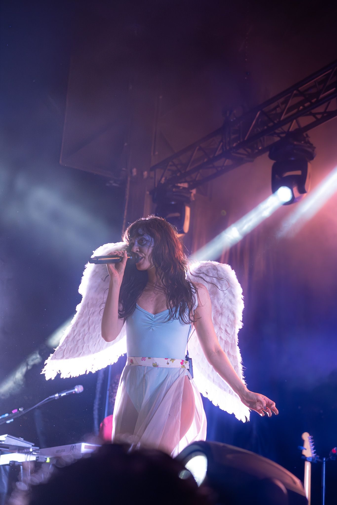 Female singer in angel wings costume performs on stage with dramatic lighting and smoke effects.