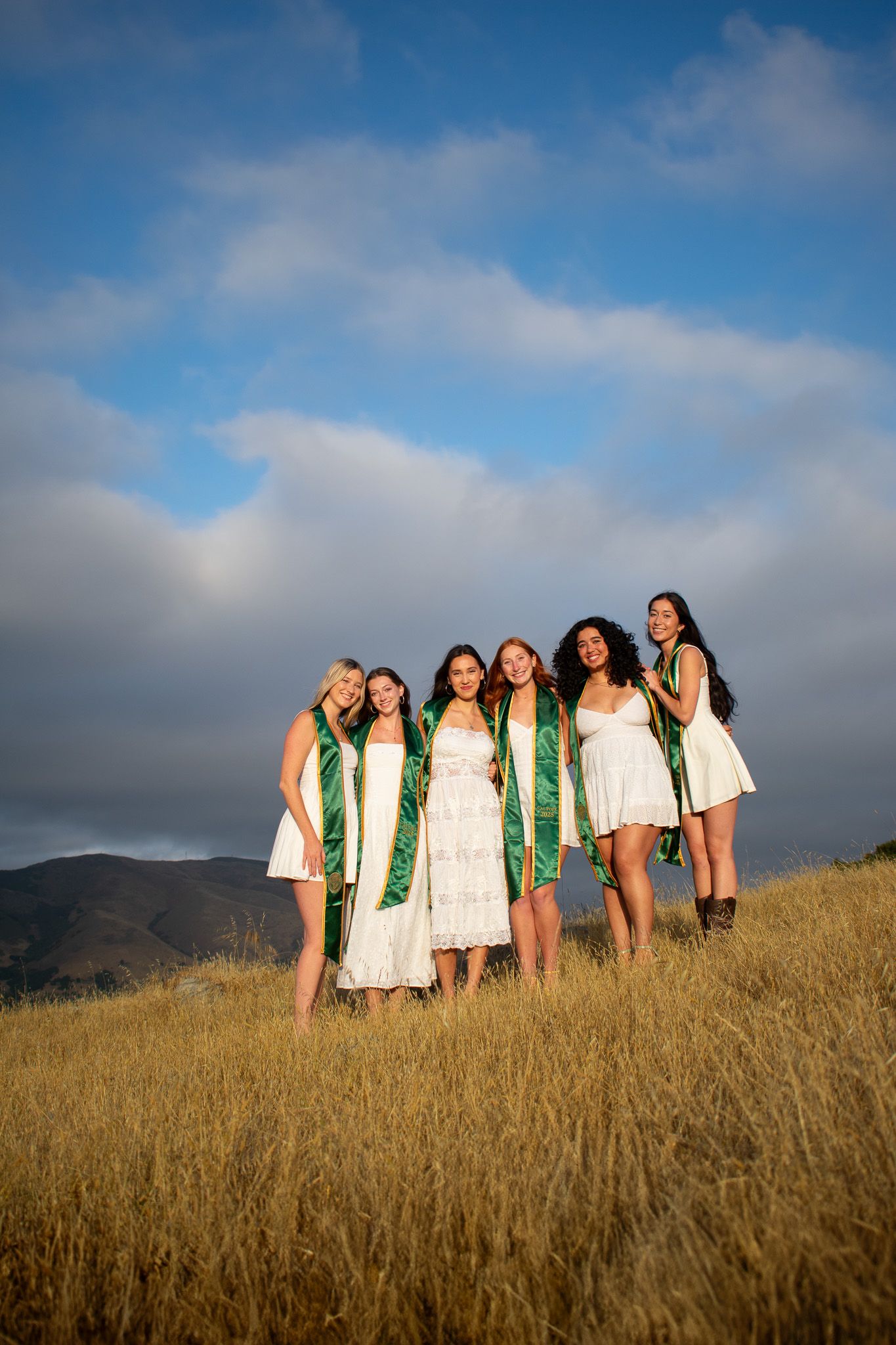 Group photo of graduates in white dresses with green stoles standing in golden grass field at sunset.