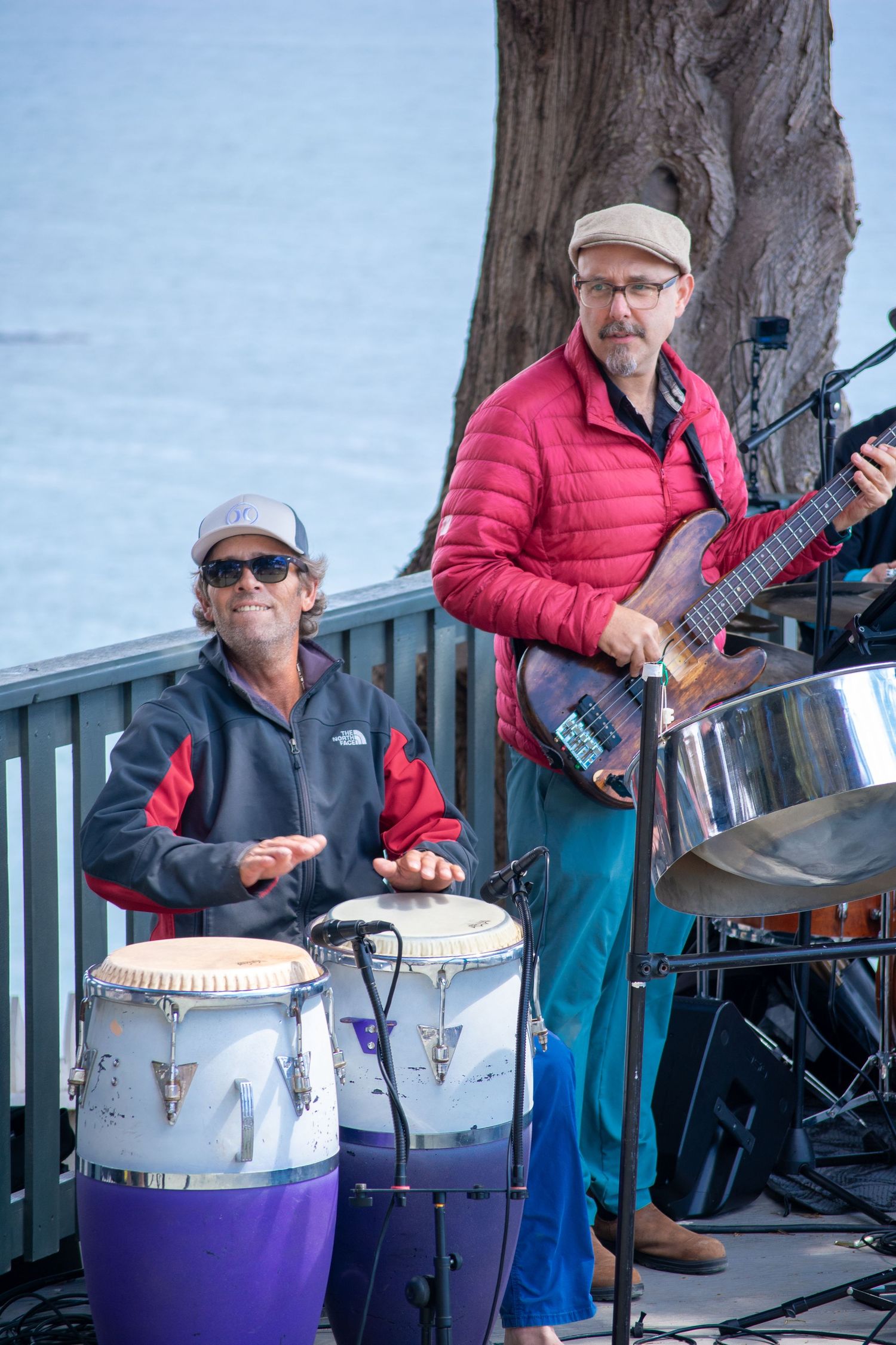 Band members perform with drums and instruments at outdoor waterfront venue.