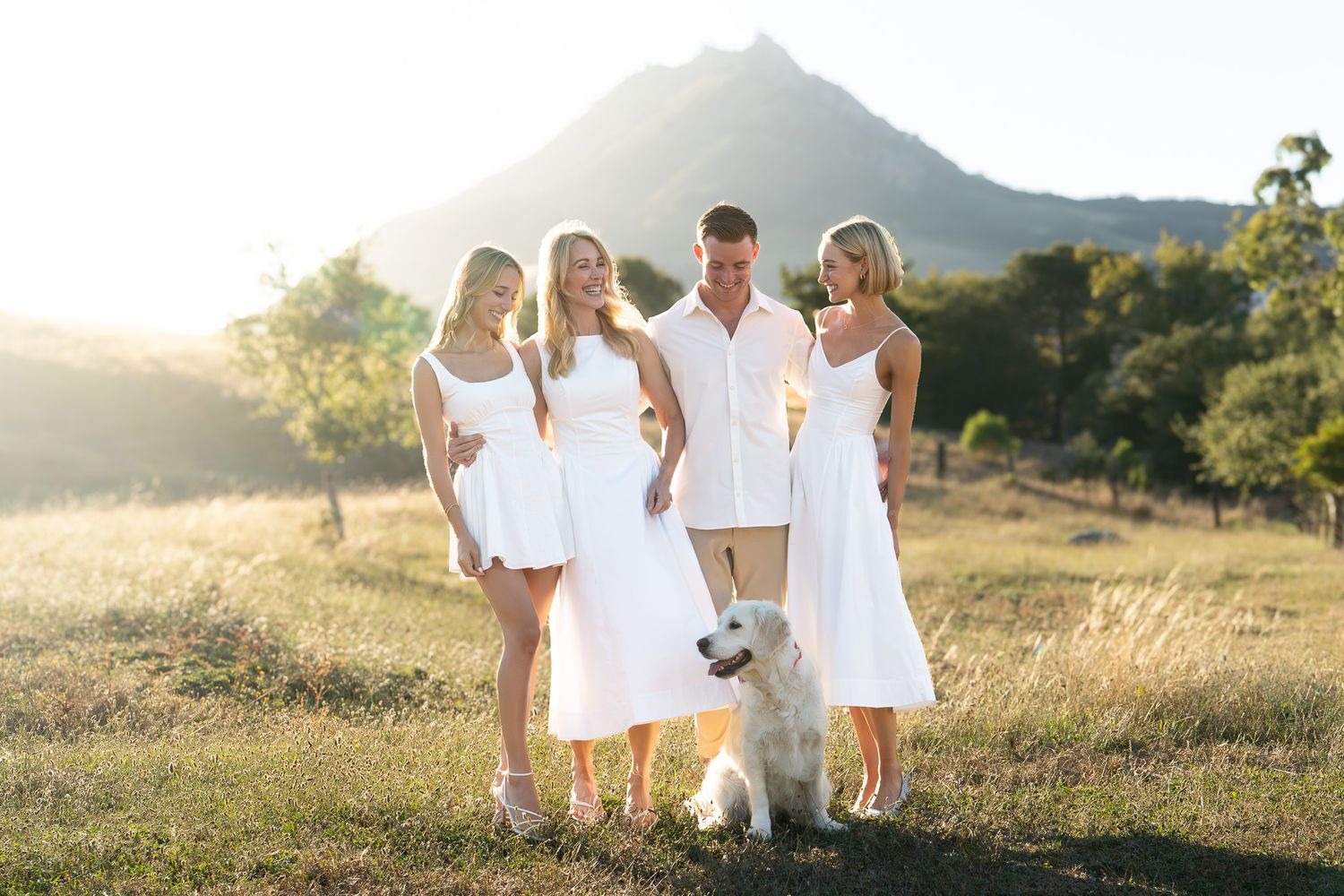 Family of three women and a man in white outfits pose with a golden retriever in a sunny field with a mountain backdrop.