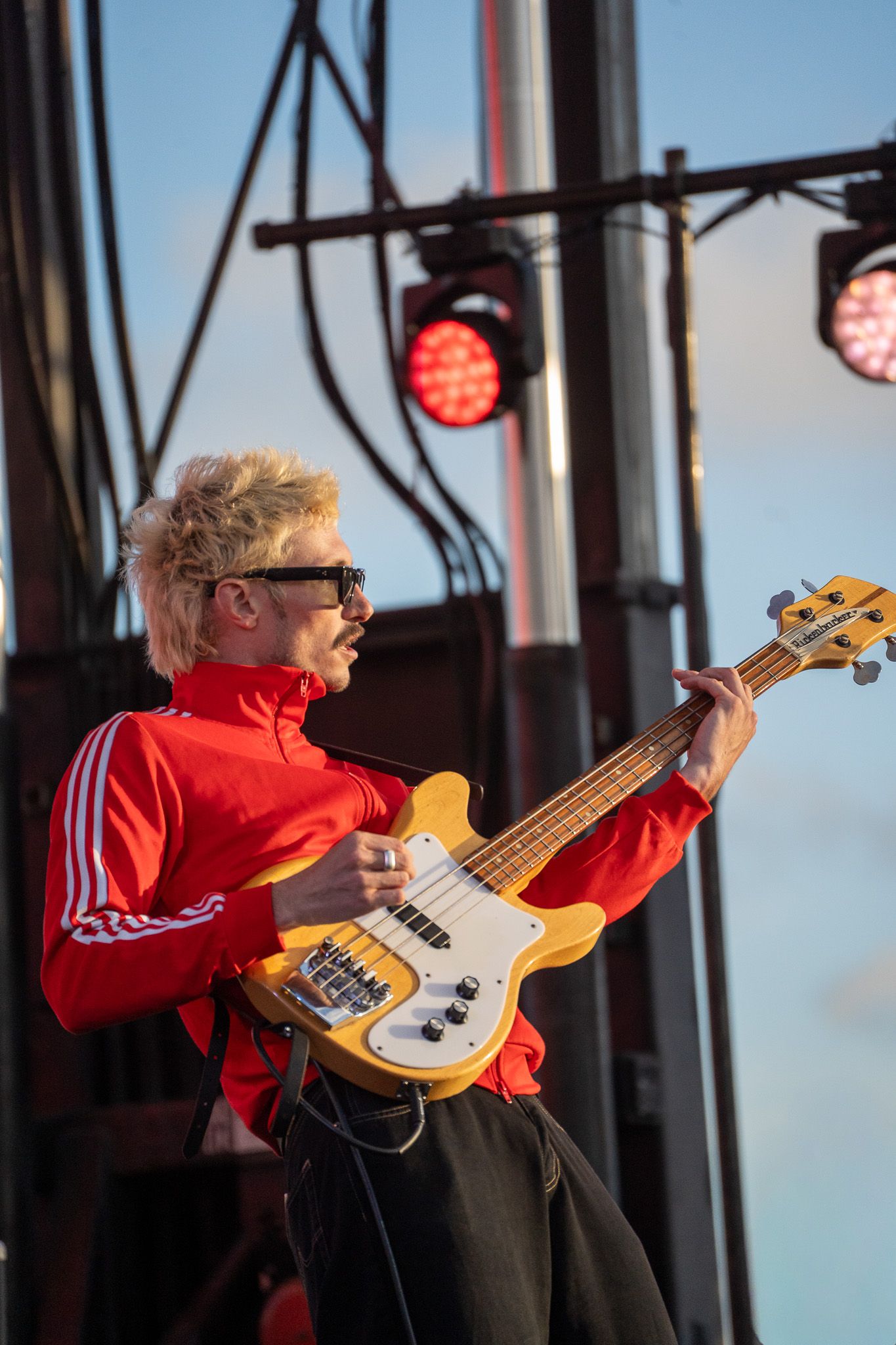 Blonde guitarist in red track jacket plays yellow bass guitar on outdoor stage with red stage lights.