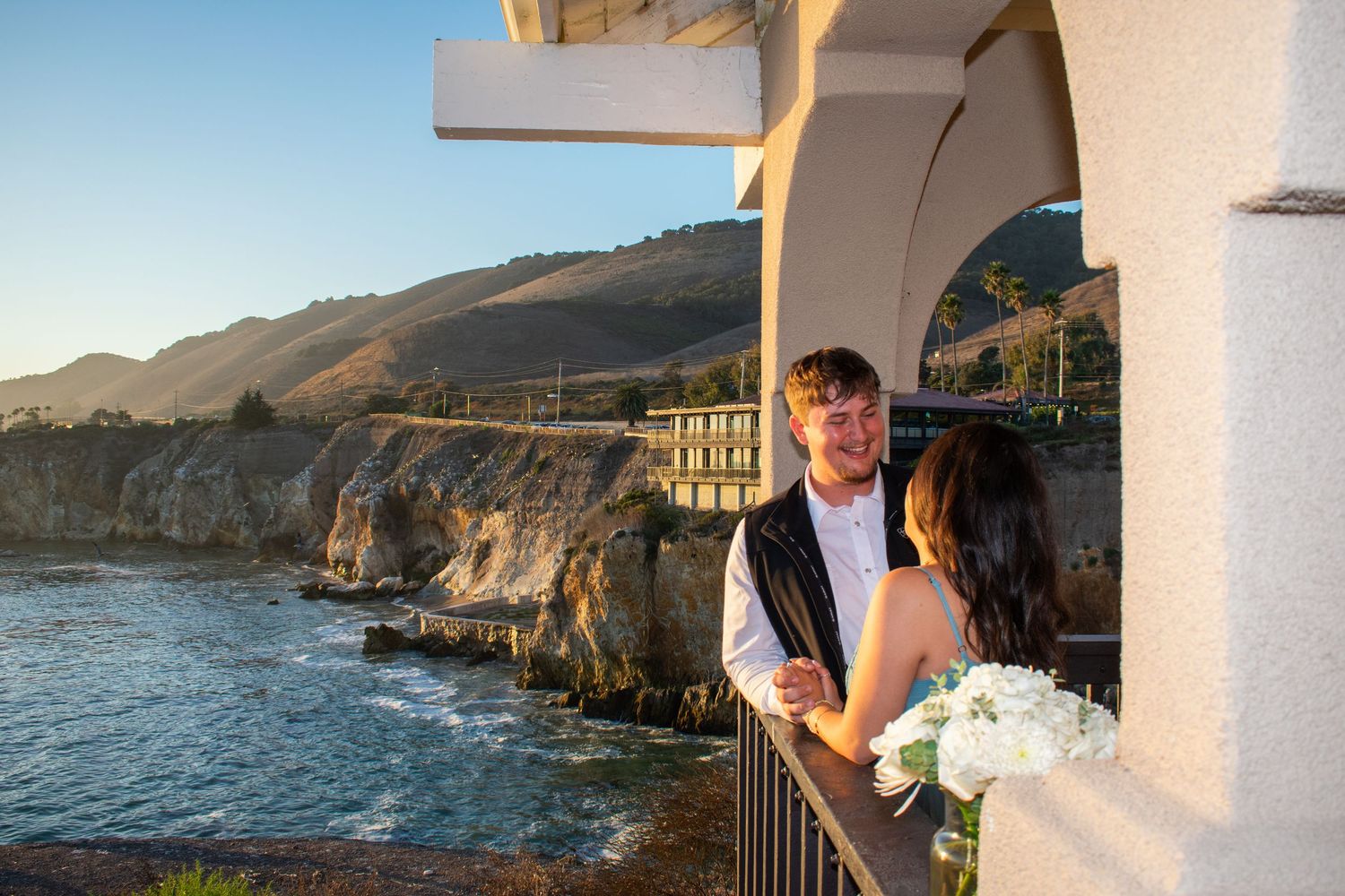 A romantic sunset proposal moment on a coastal balcony overlooking dramatic cliffs and ocean views.