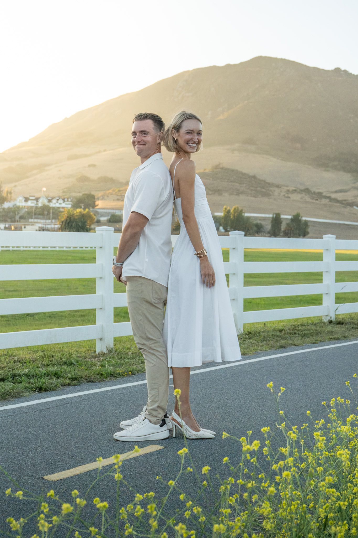 Couple in white outfits stand back-to-back on scenic road with white fence and mountain backdrop.