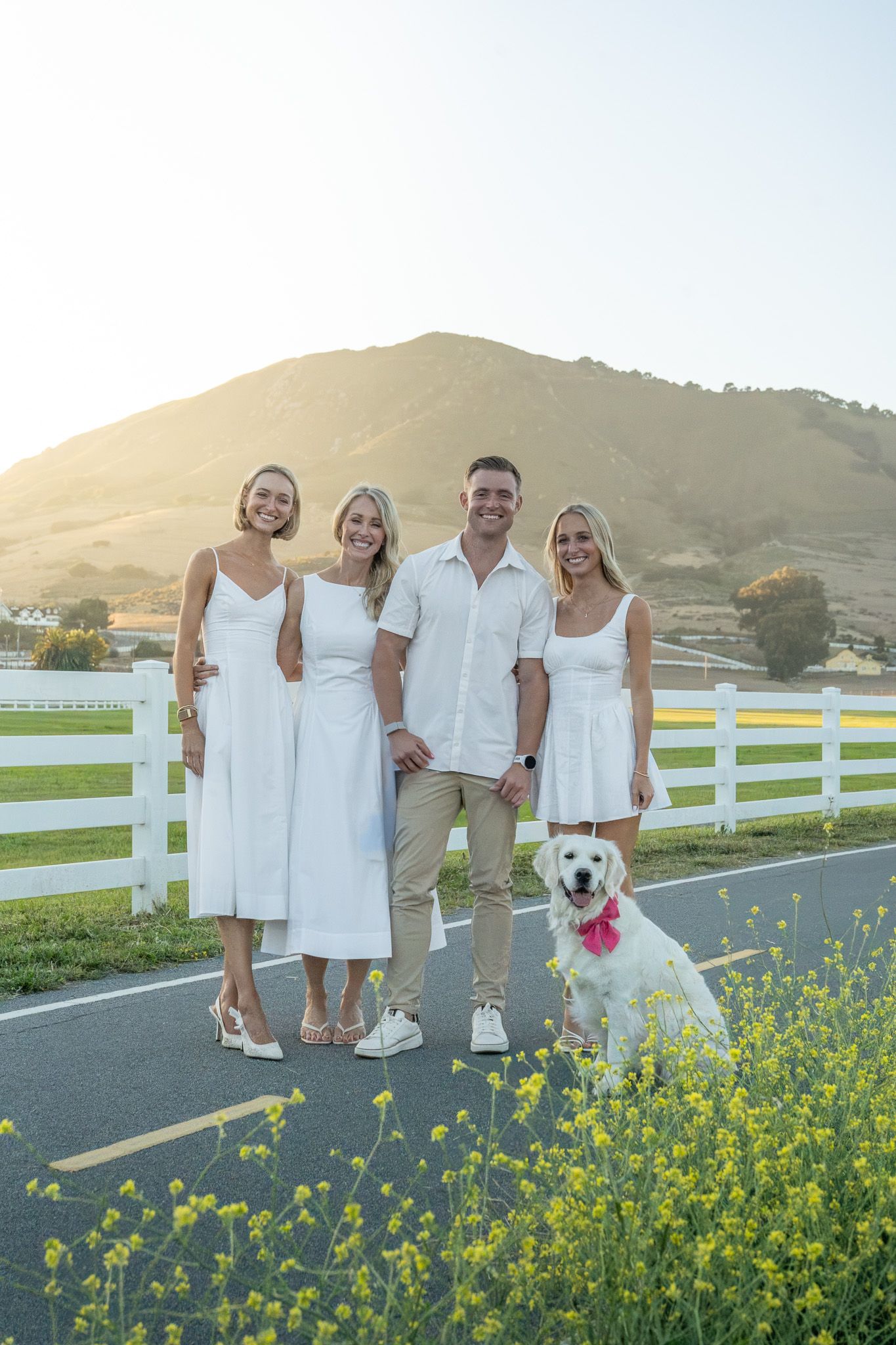 Family of four in white outfits posing with a white golden retriever by a fence, hills and sunset in background.