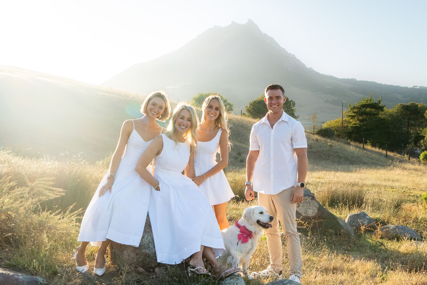 Three women in white dresses and a man with a dog pose on a sunny hillside with a mountain backdrop.
