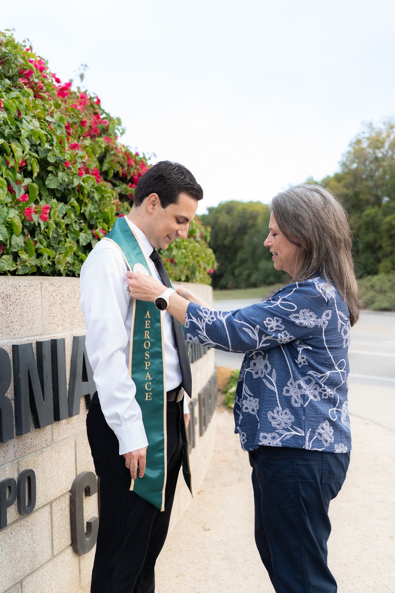 Woman adjusting green Aerospace graduation sash on man near Cal Poly campus sign with red flowers.