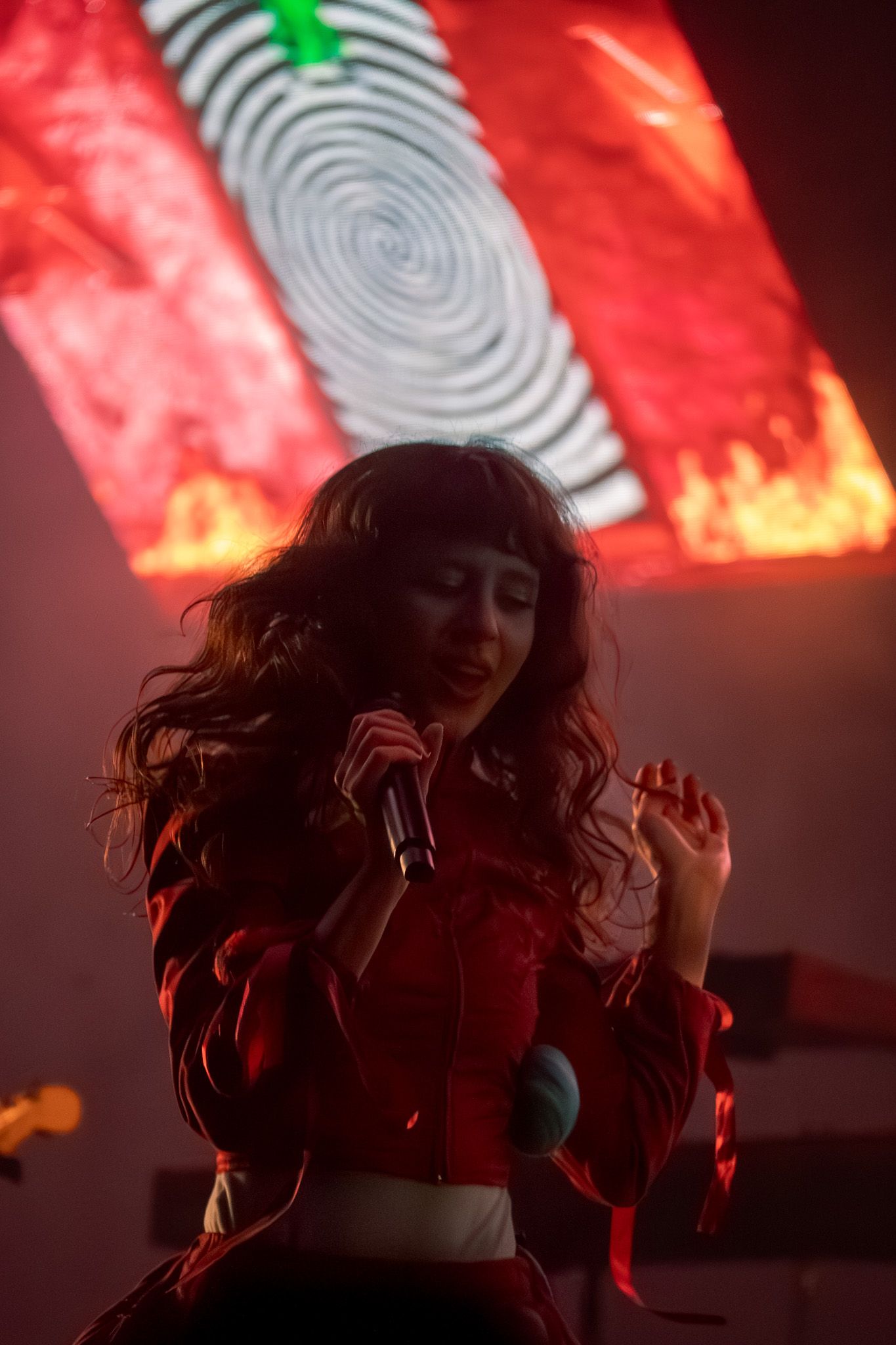 Female singer in red outfit performs on stage holding microphone with vibrant red spiral projection backdrop.