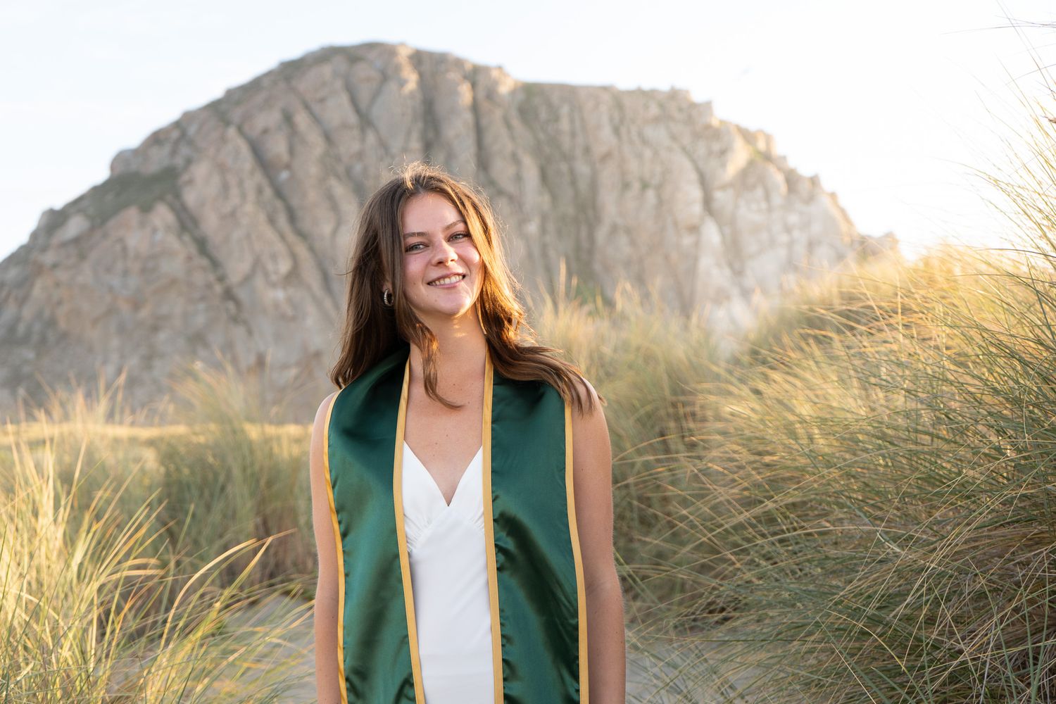 Young woman in green graduation regalia smiles on sandy dunes with Morro Rock in the background.