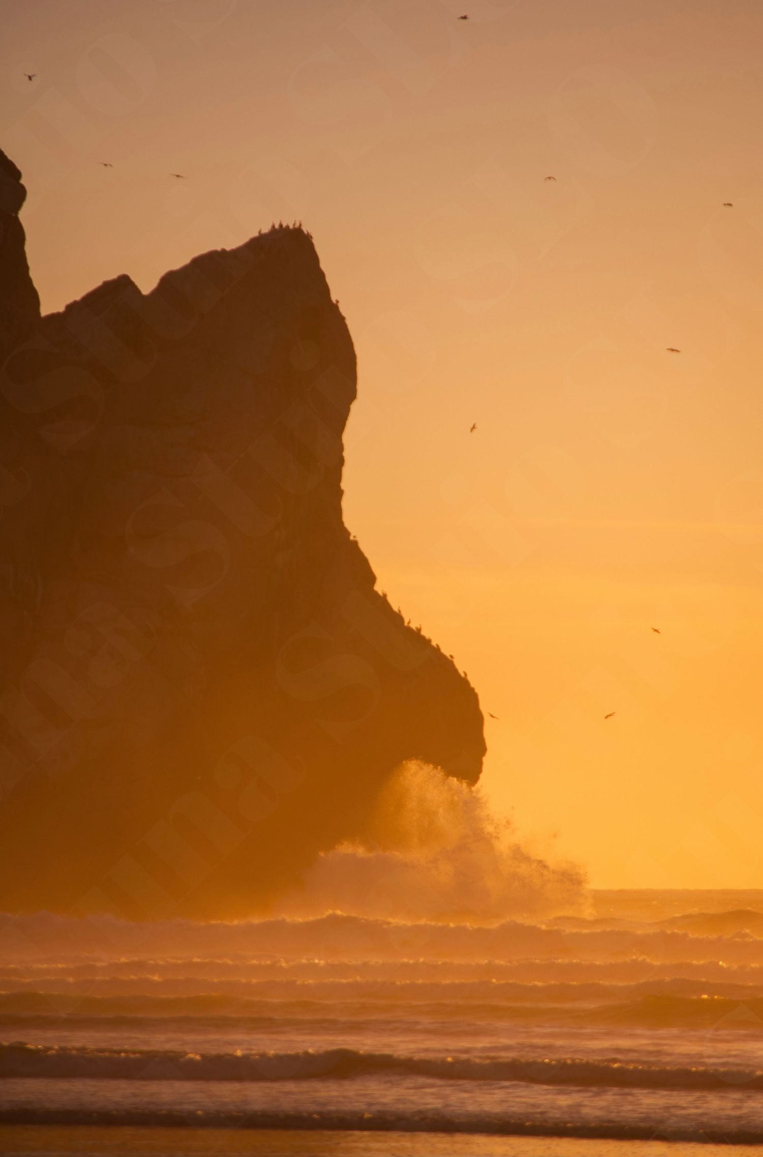 Dramatic coastal rock formation silhouetted against an orange sunset sky with crashing waves.