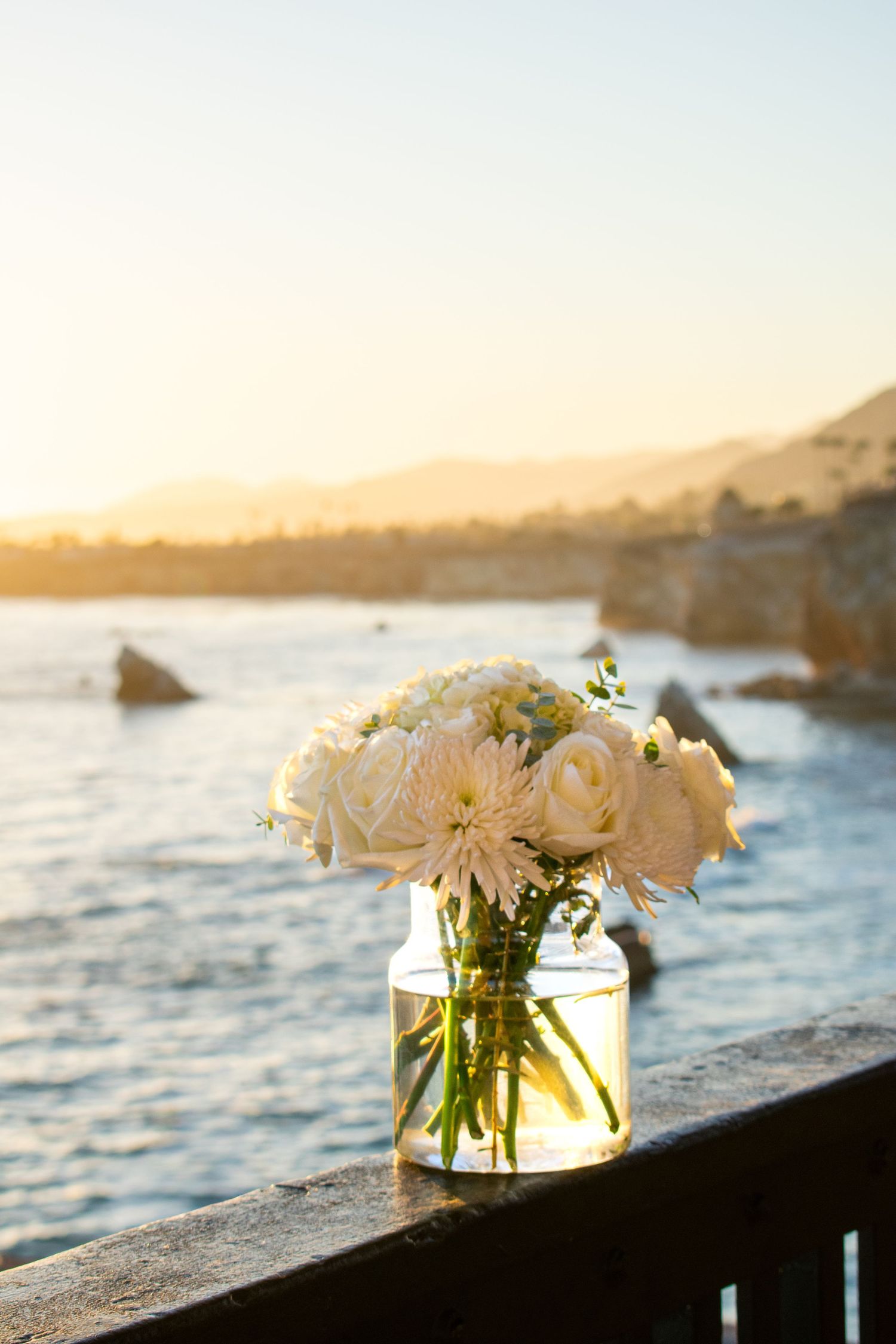 Glass vases filled with white flowers displayed on stone wall with sunset ocean view.