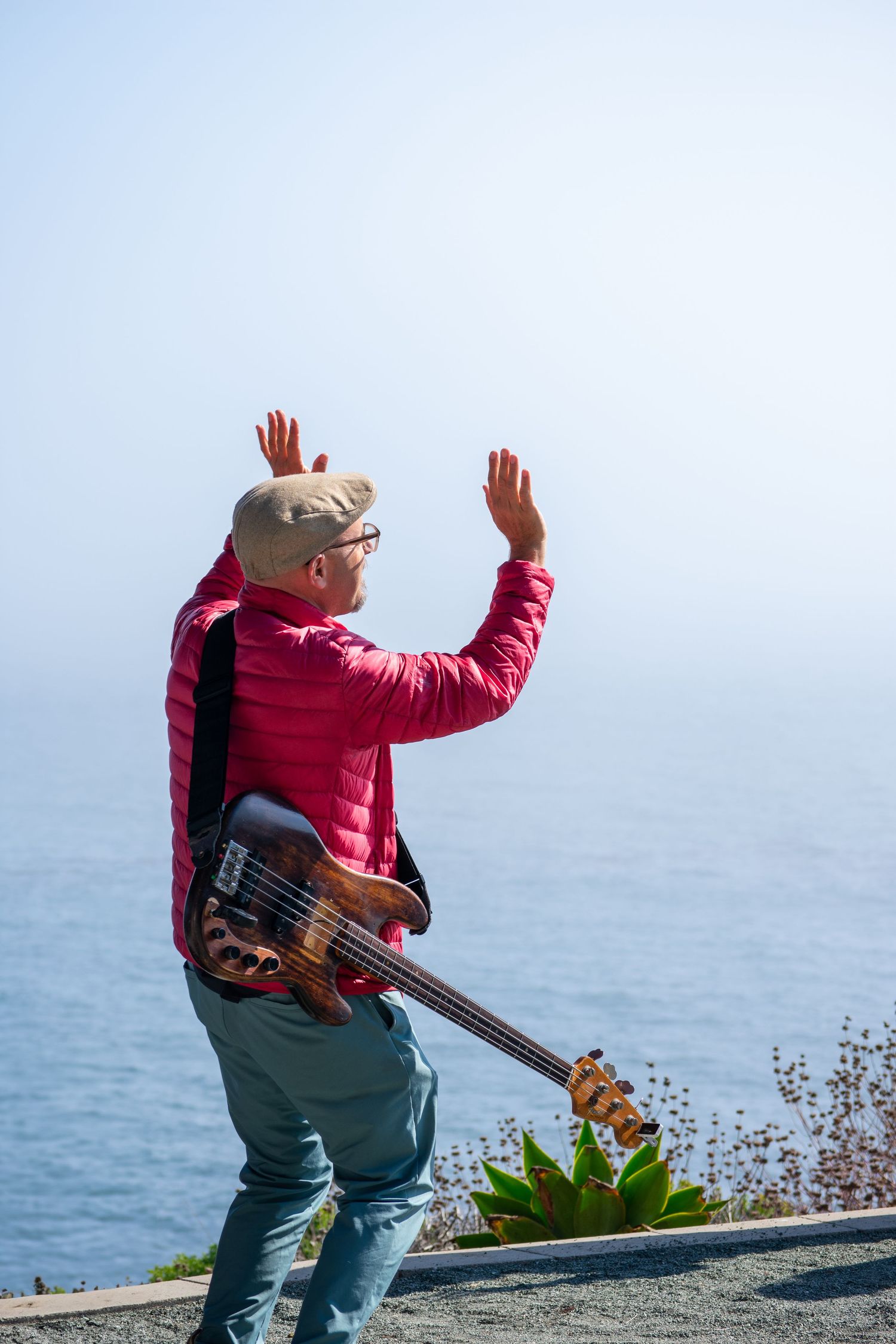 Bass guitarist in red jacket performs against ocean backdrop with raised arms.