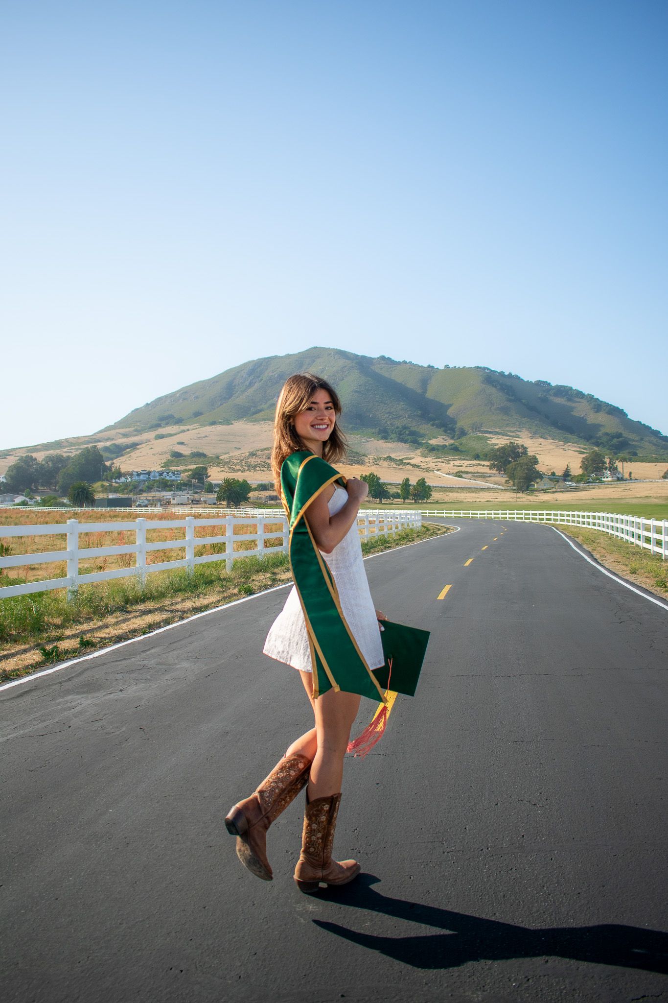 Graduate in green stole poses playfully on empty road with mountain backdrop.