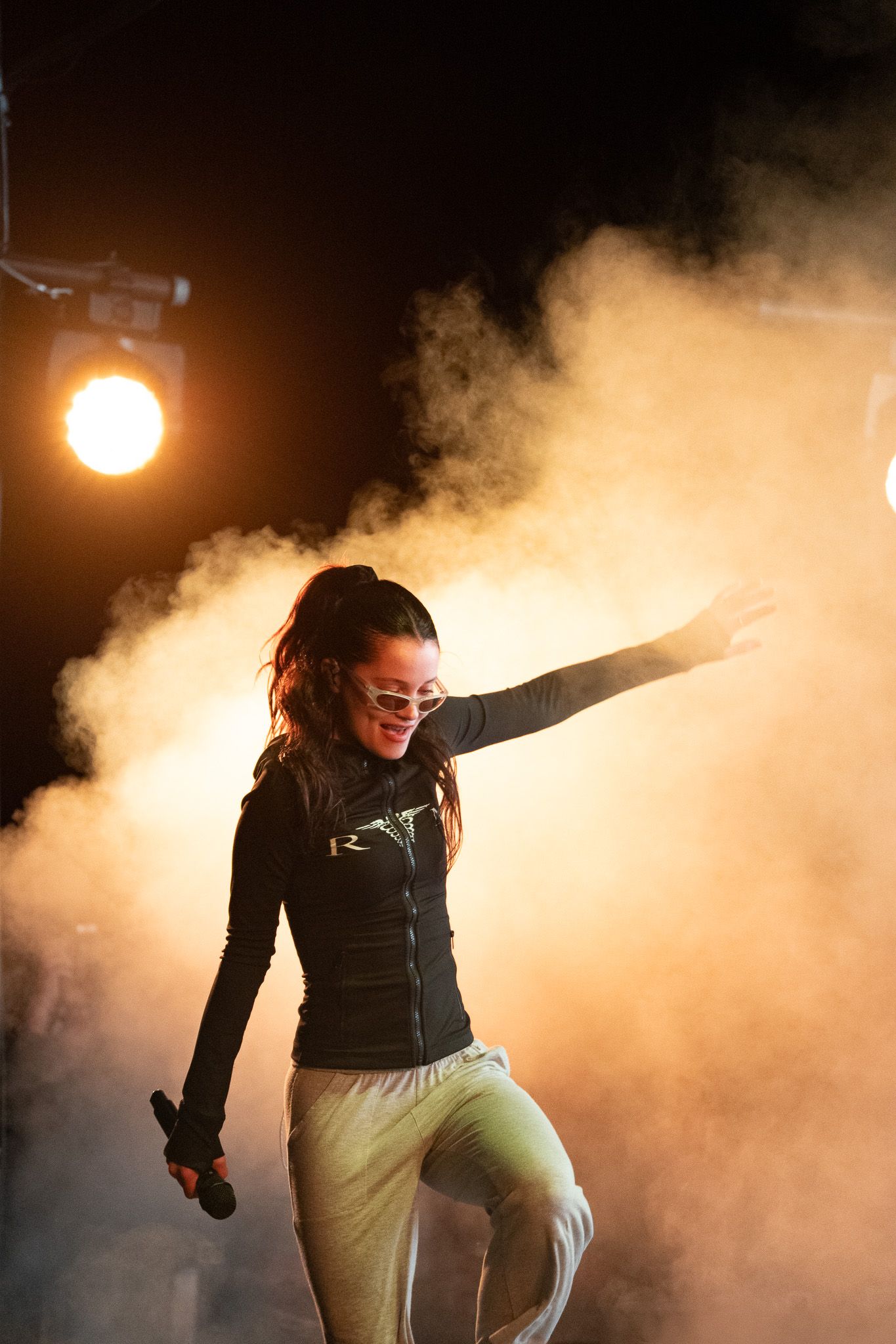 Female performer holds microphone on smoky stage, arms outstretched, wearing sunglasses and black jacket under dramatic spotlight.