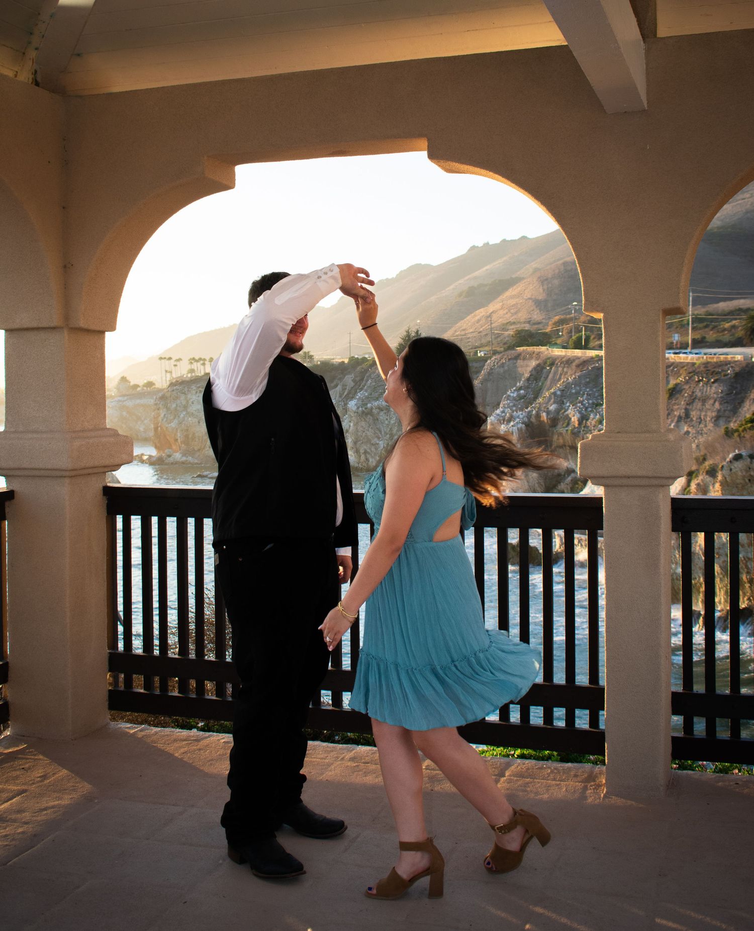 Dancing couple in formal attire on a covered balcony with ocean and mountain views.