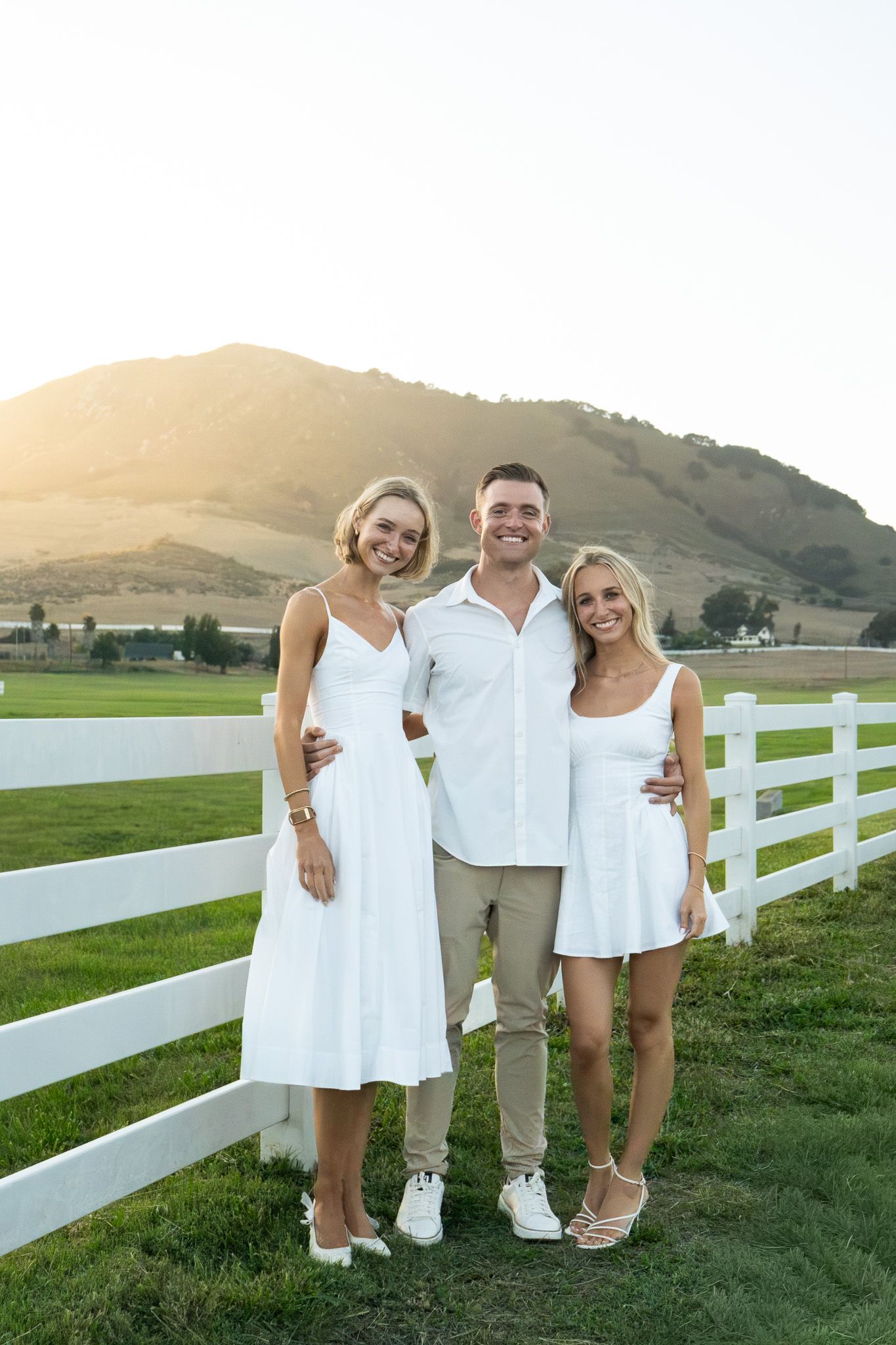 Three friends in white outfits smile together by a white fence with a sunlit mountain backdrop.