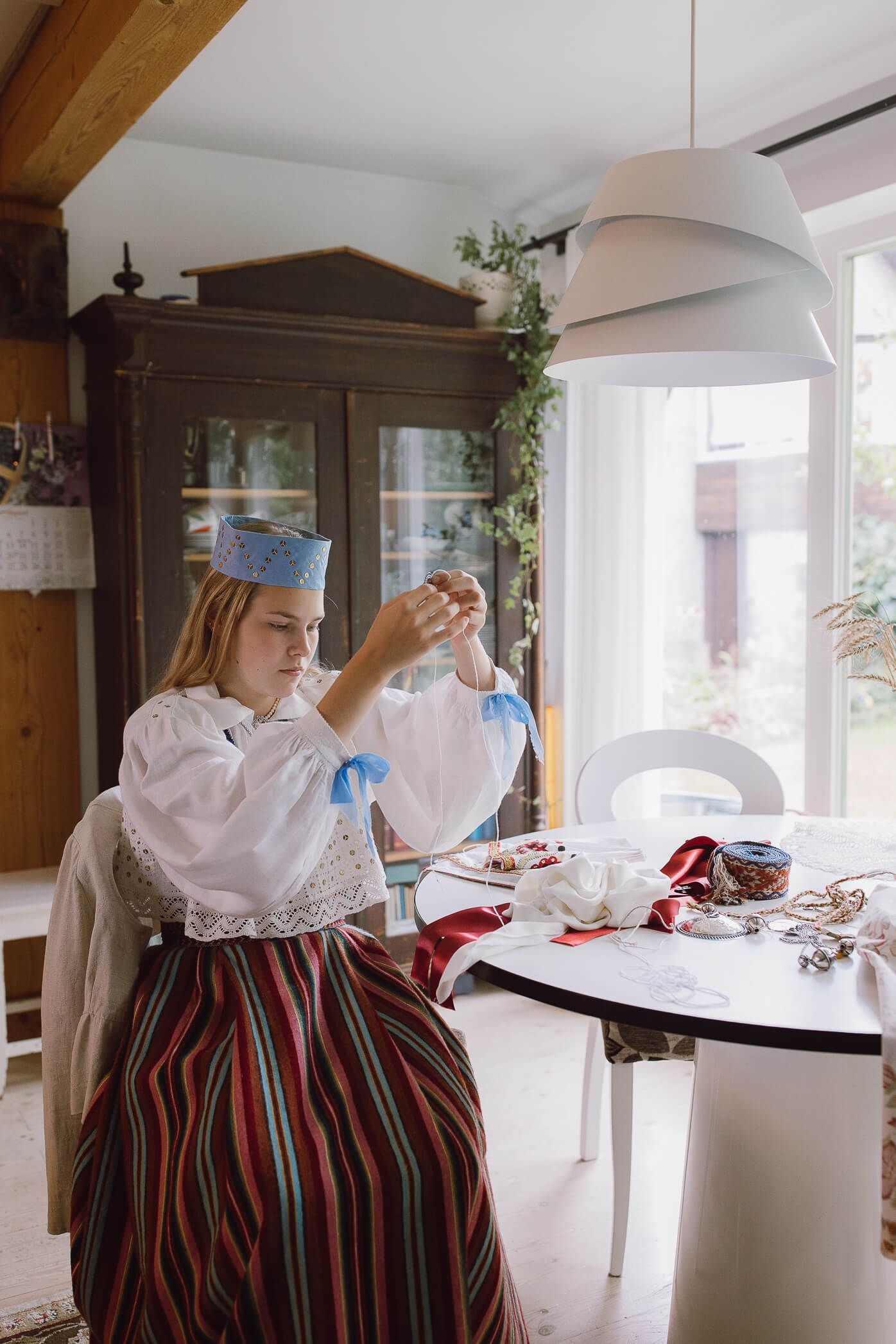 Young woman in traditional folk costume with striped skirt and blue crown hat arranging jewelry at a dining table.