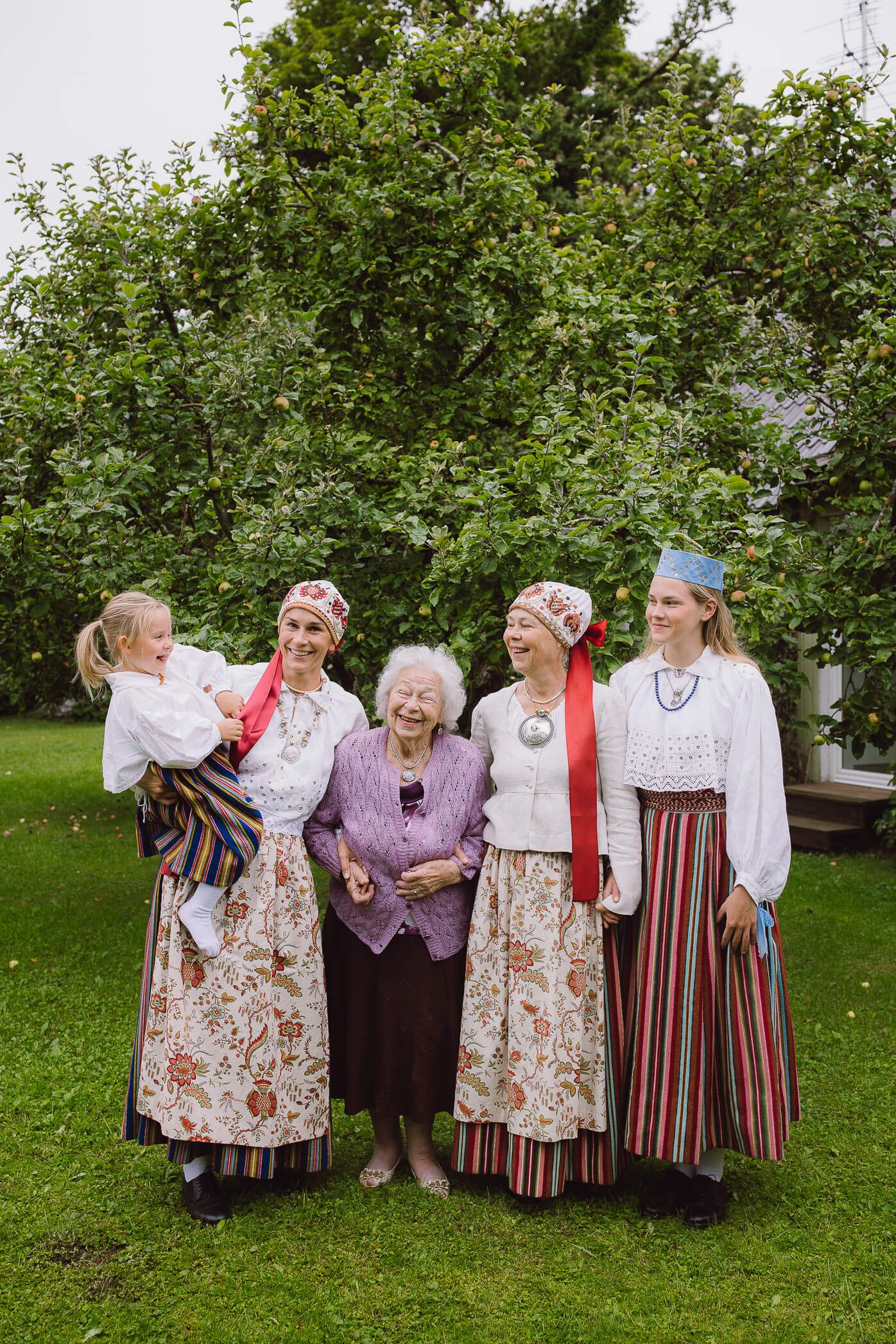 Five women in traditional folk costumes smile together outdoors, with lush green trees in the background.
