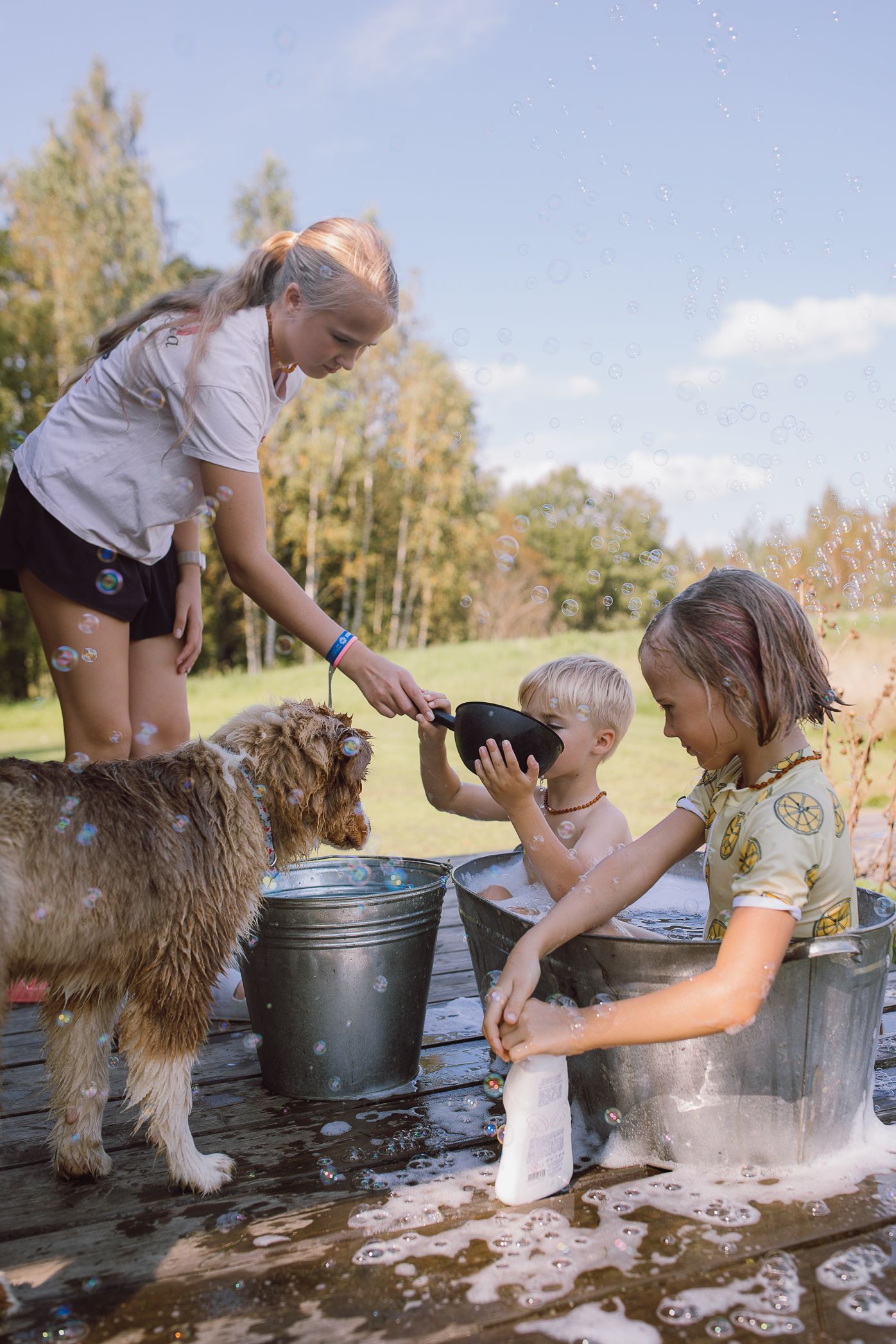 Children and a fluffy dog gathered around metal buckets outdoors on a sunny day.