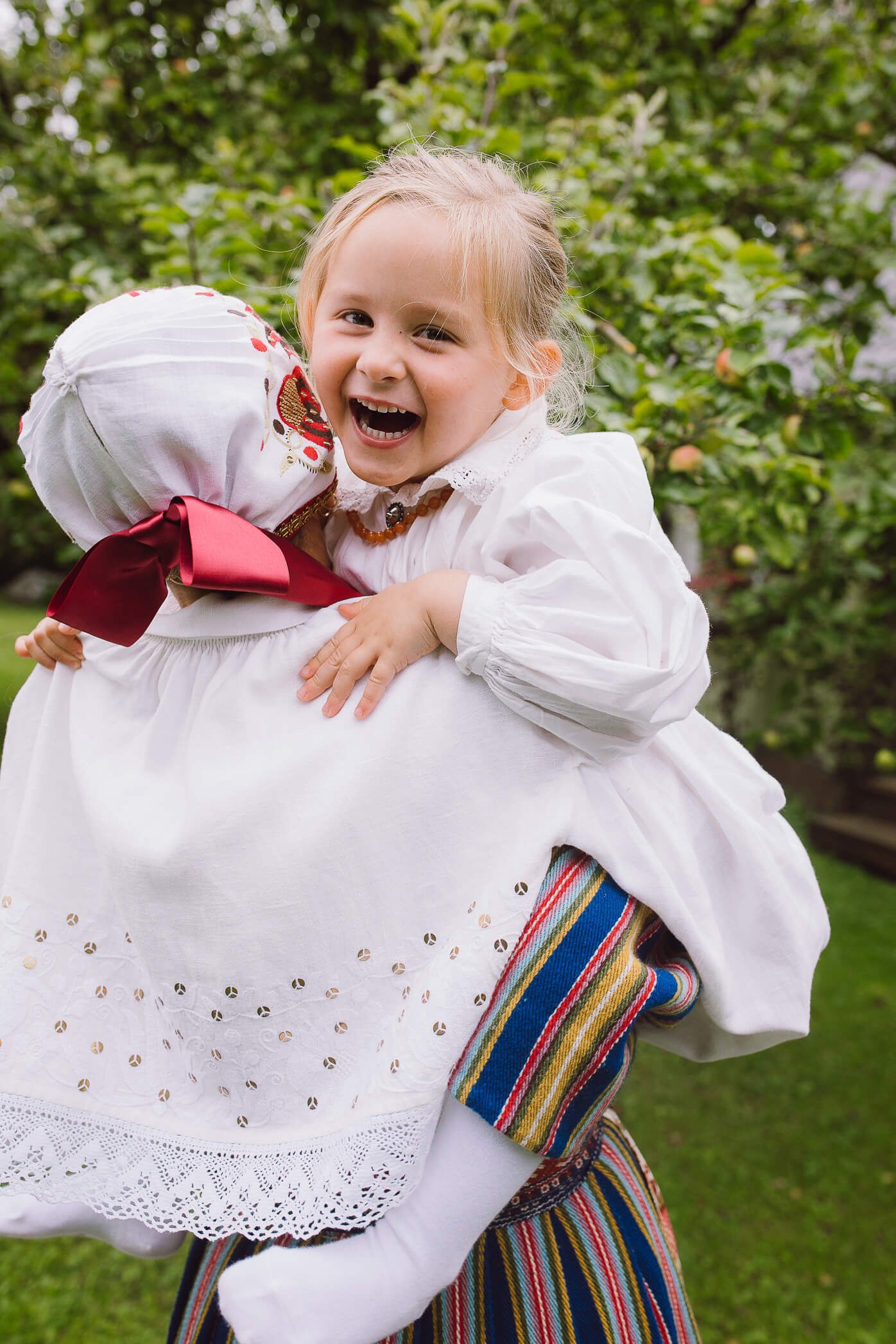 A joyful child in traditional folk costume being held by an adult in matching attire outdoors.