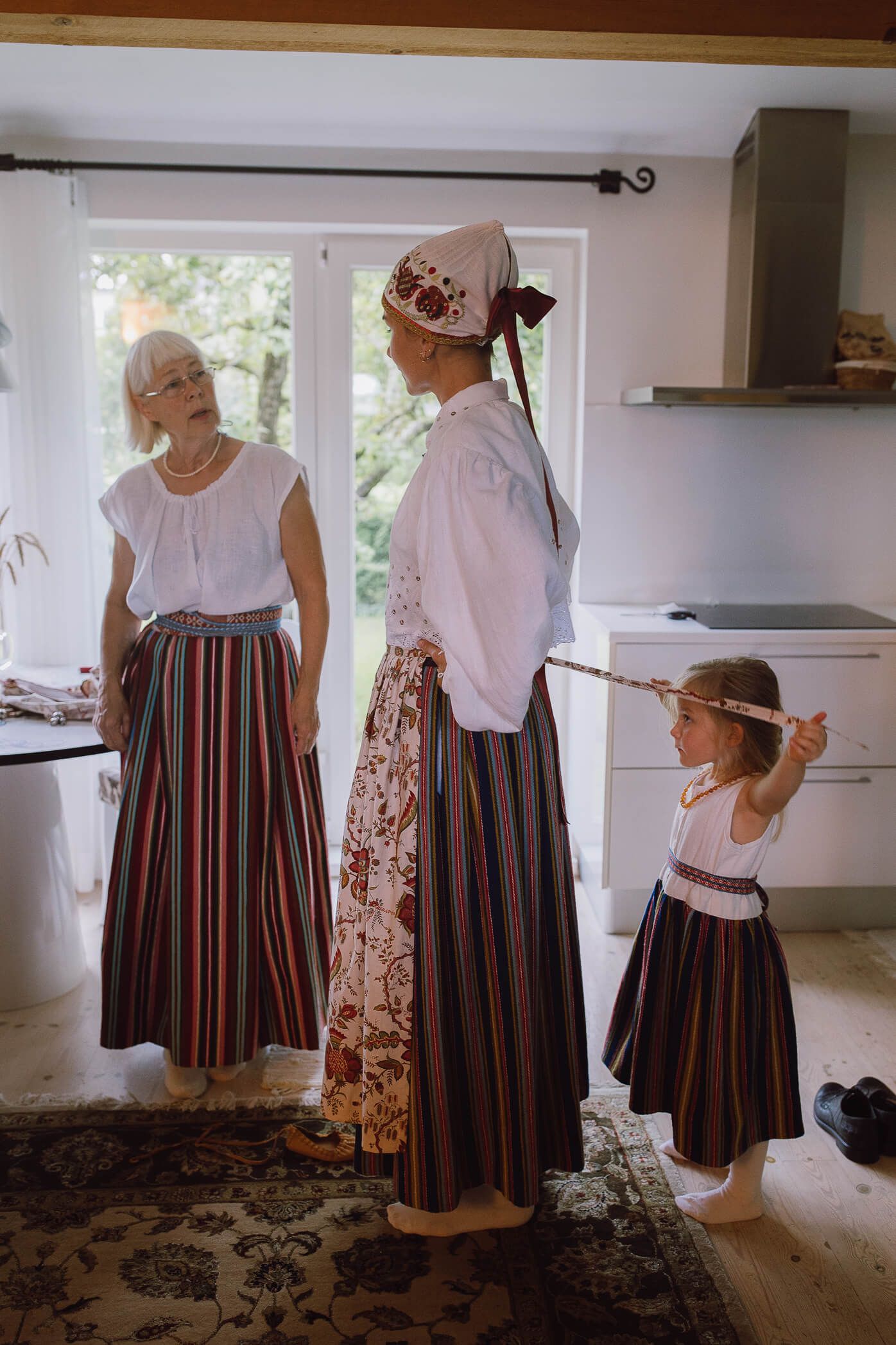 Three generations of women in traditional Estonian folk costumes with striped skirts standing together indoors.
