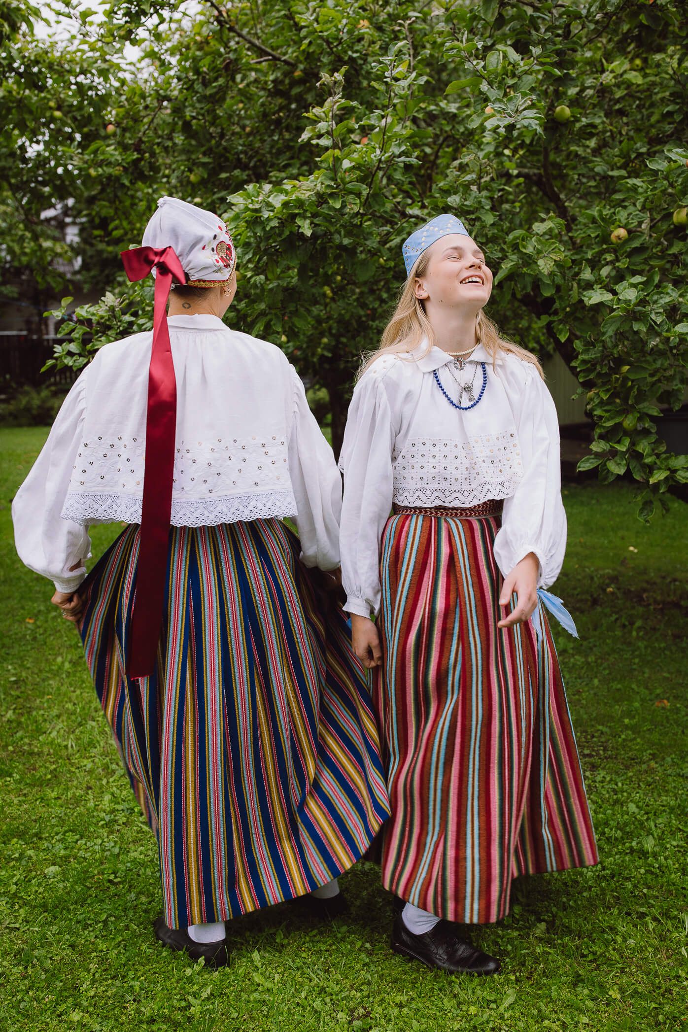 Two young women in traditional Nordic folk costumes with striped skirts and white blouses stand outdoors laughing.