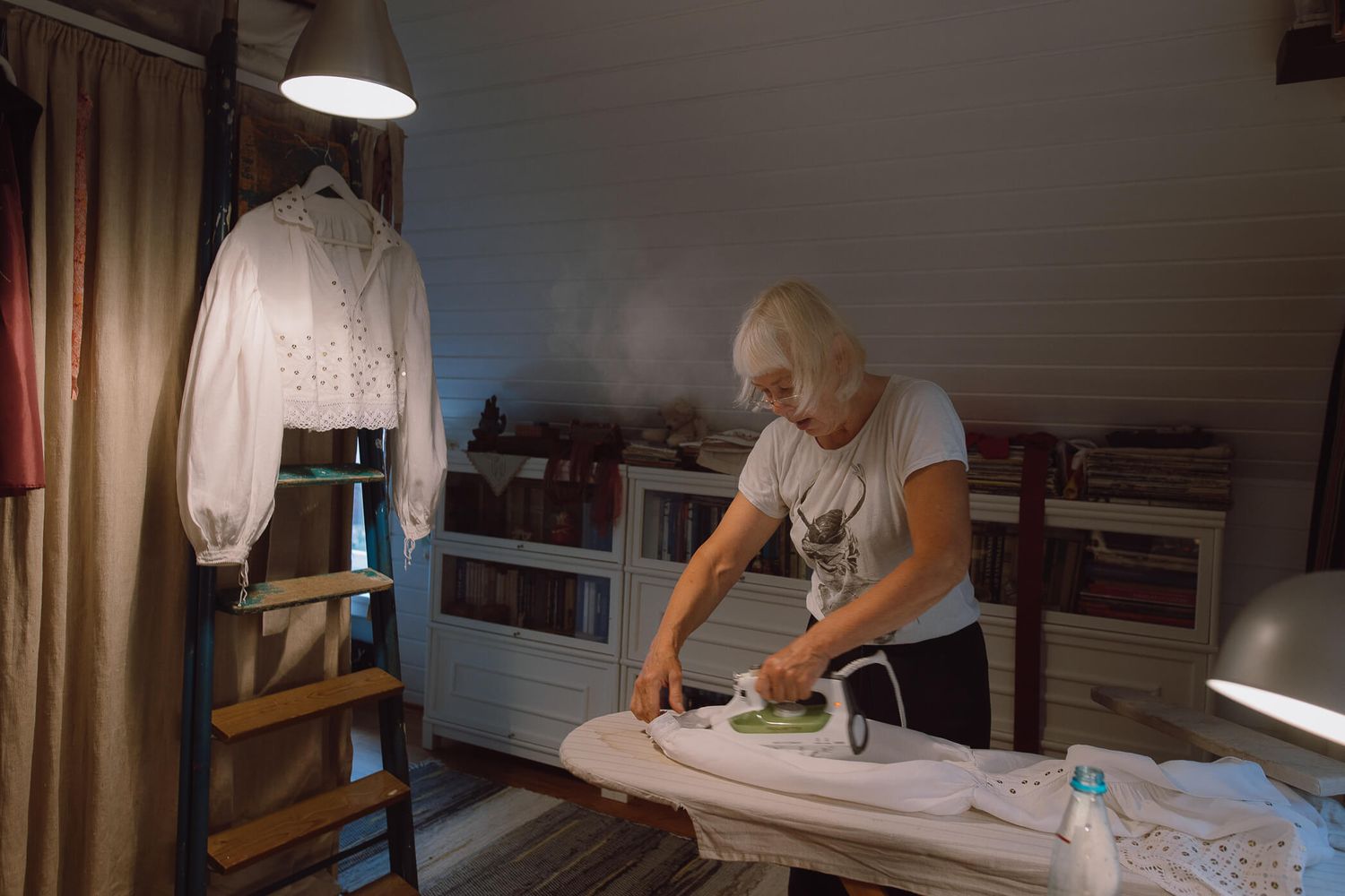 Elderly woman ironing clothes on a board in a cozy room with a white blouse on a ladder stand nearby.