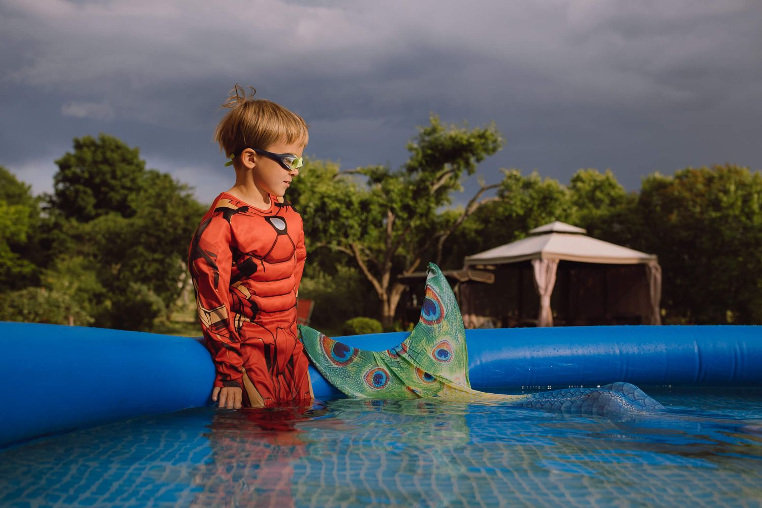 Child in orange swimwear plays in a blue inflatable pool under stormy skies.