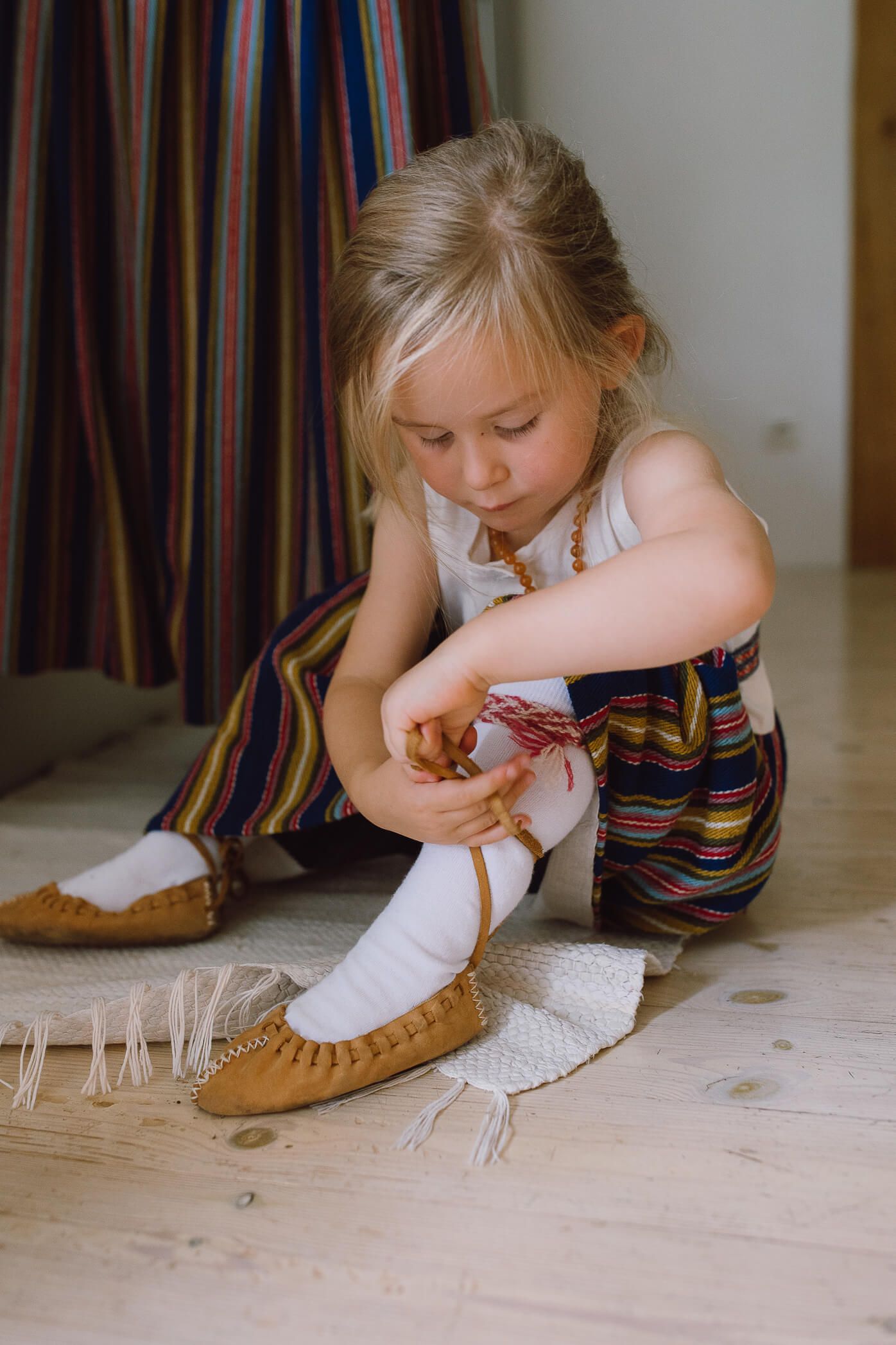 Young blonde girl in colorful striped outfit sitting on floor tying laces on brown moccasin shoes.