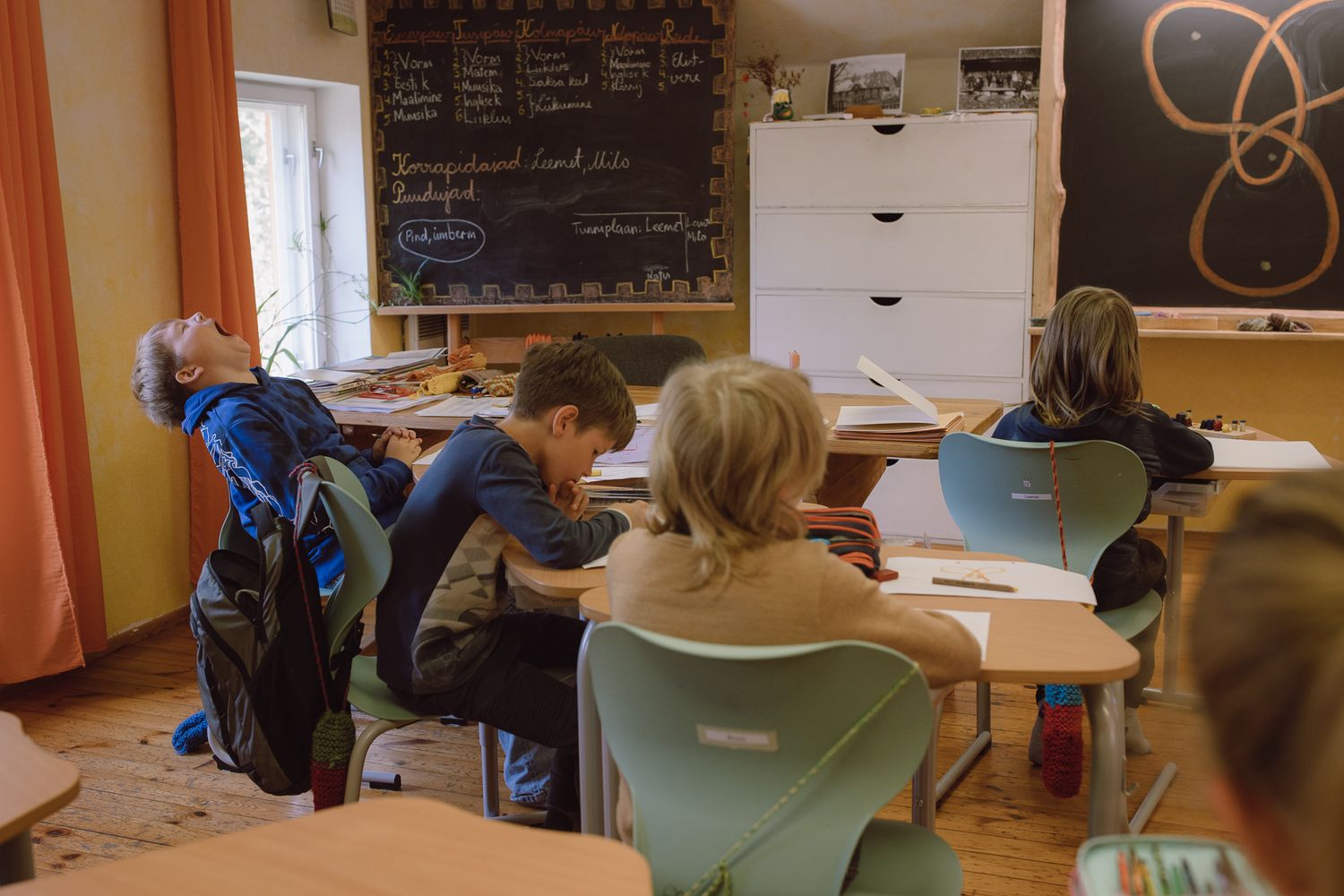 Students seated at desks while teacher writes on chalkboard in traditional classroom.