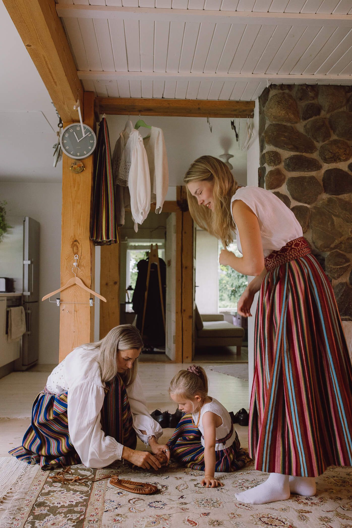Two women in traditional folk costumes help a young child dress on a rug inside a rustic home.