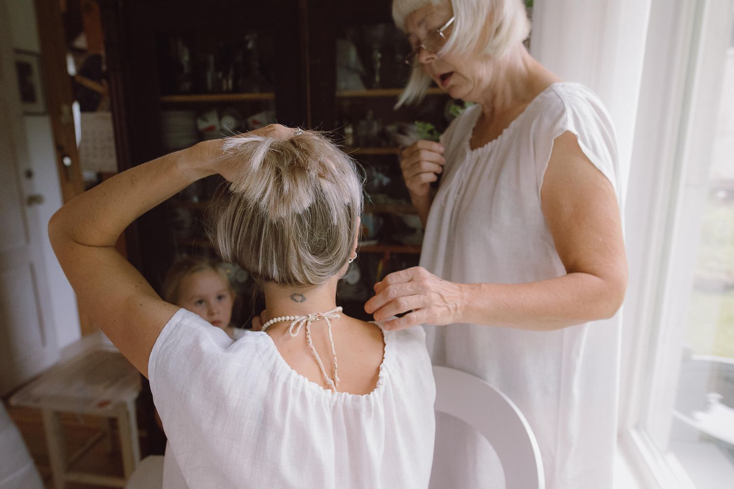 An older woman helping a younger woman with her hair in a warmly lit, vintage-style room.