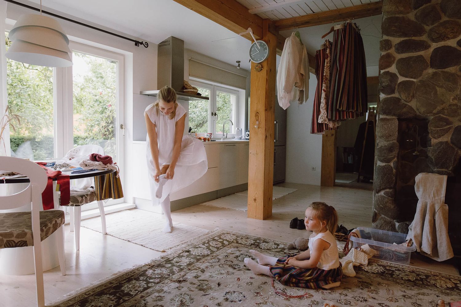A woman in a white dress adjusts her outfit while a young girl plays on a rug in a cozy Scandinavian-style home.