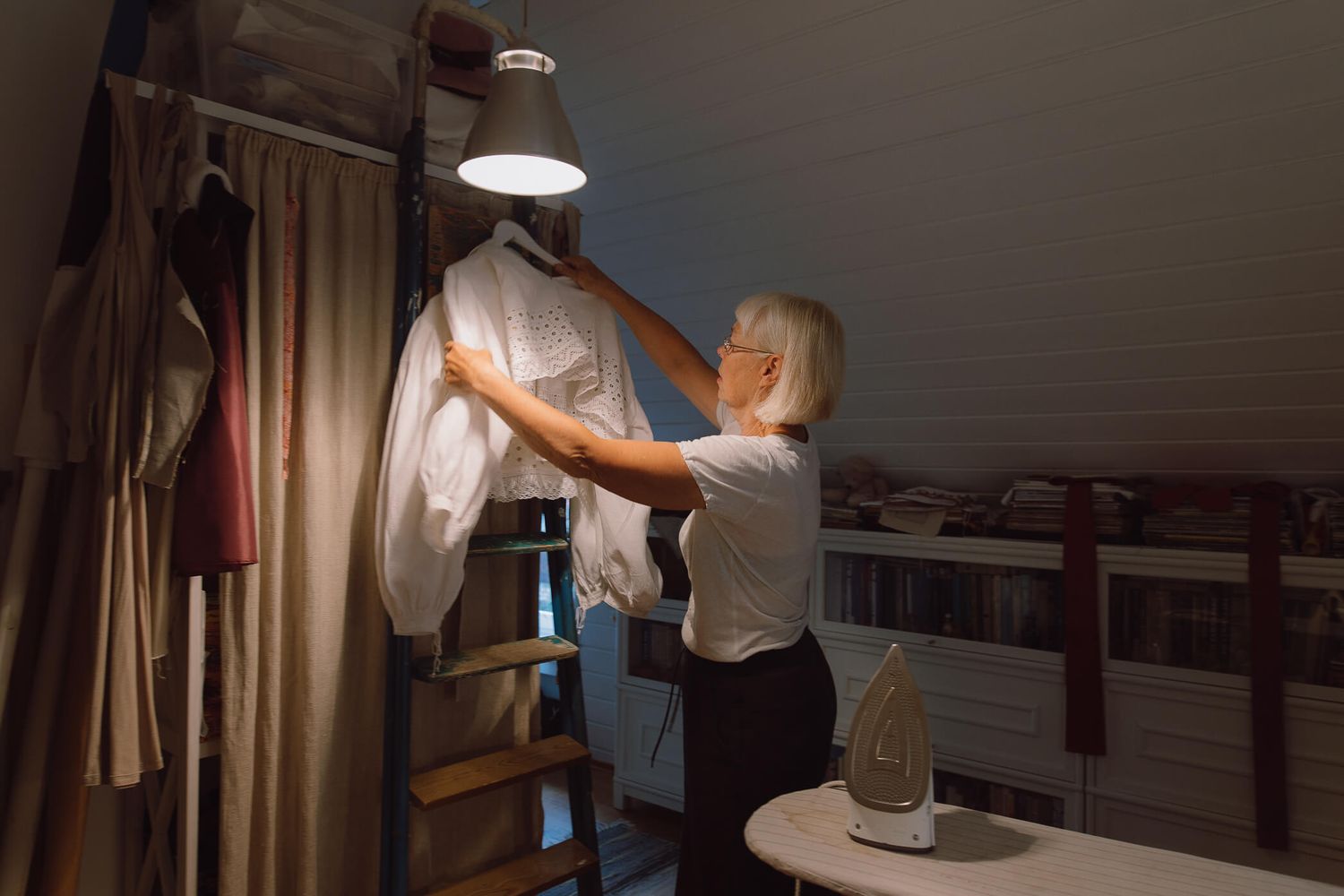 Woman organizing clothes on a rack under a pendant light in a dimly lit attic closet space.