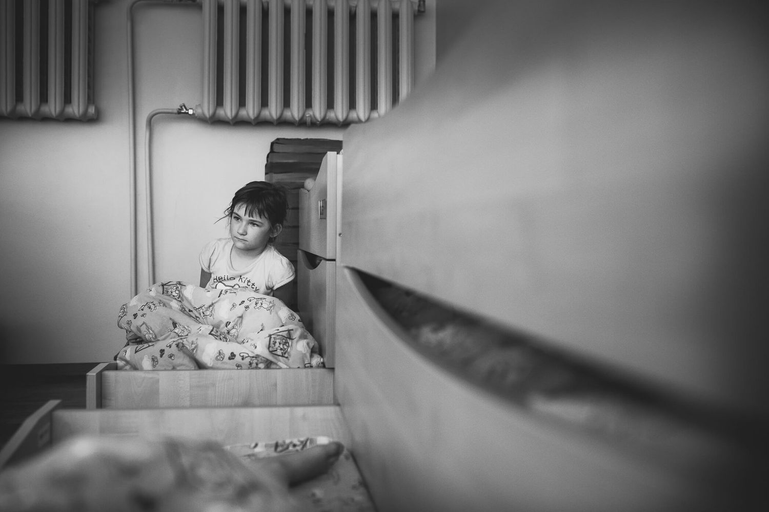A black and white photograph shows someone resting in a hospital bed with radiator pipes visible on the wall.