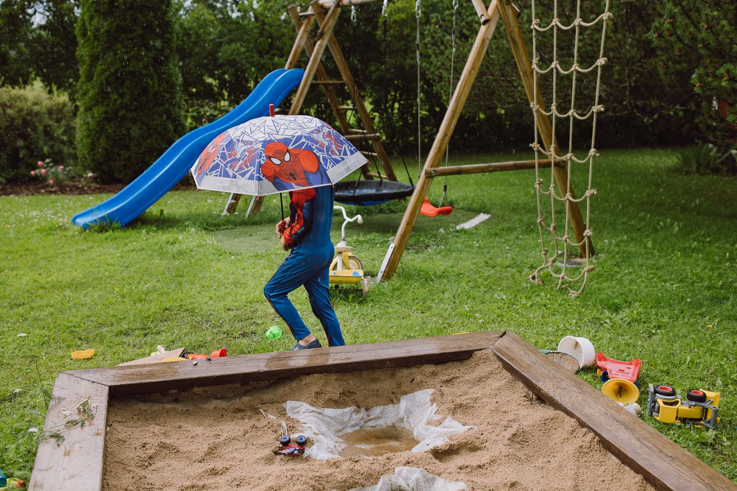 Person with umbrella jumps in a wet sandbox near playground equipment on a rainy day.