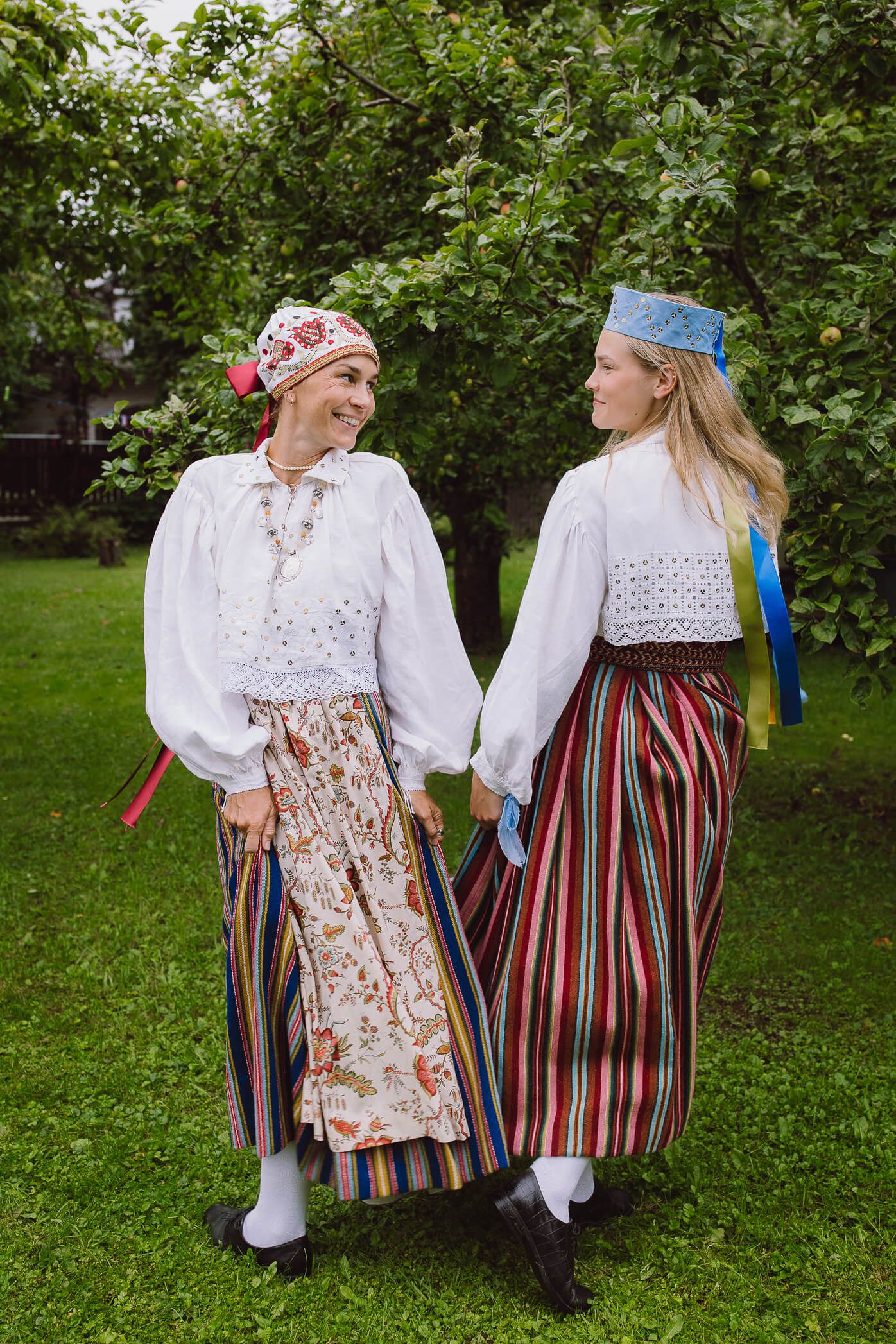 Two women in traditional Estonian folk costumes with striped skirts and embroidered blouses holding hands outdoors.