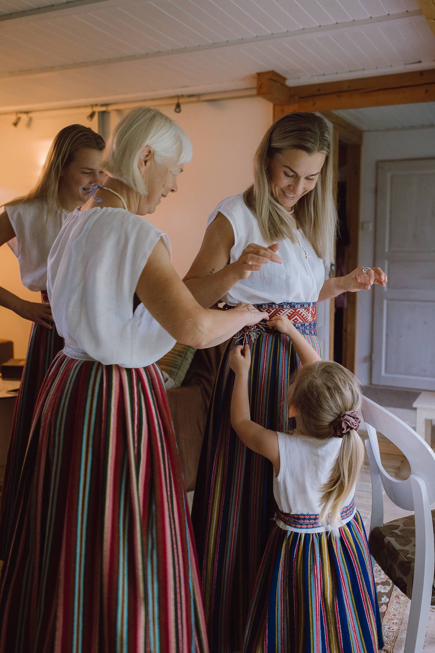 Three women and a girl help each other dress in colorful traditional folk costumes with striped skirts.